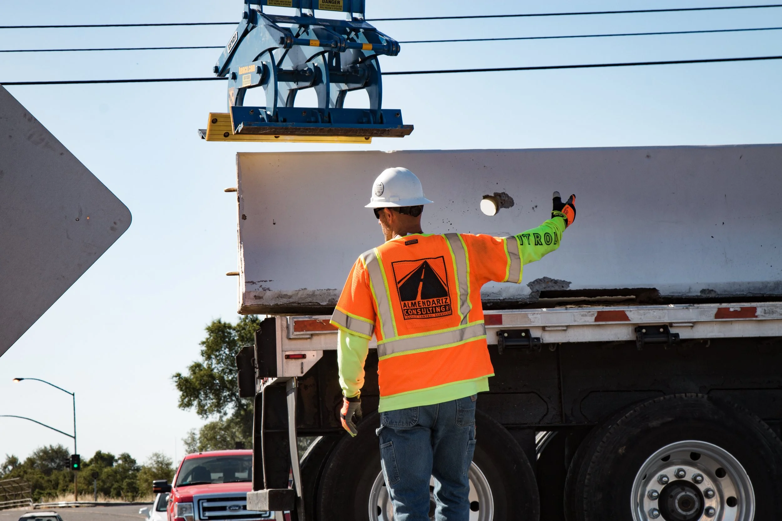 A construction worker wearing a white helmet and orange safety vest with yellow reflective stripes is directing traffic at a construction site. He is standing near a large white utility truck and pointing to the right. There are power lines and a traffic light in the background.