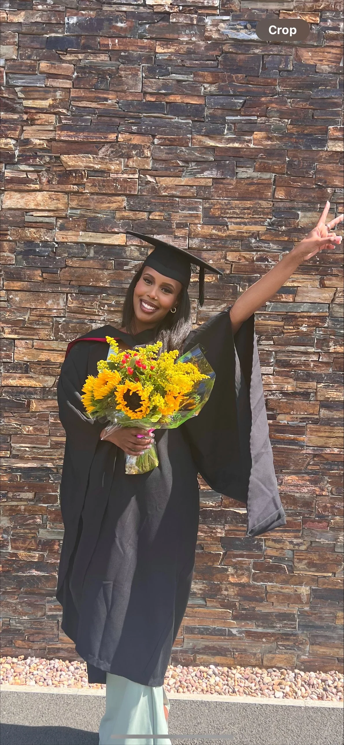 A woman in a graduation cap and gown holding a bouquet of sunflowers and yellow flowers, celebrating her graduation next to a brick wall.