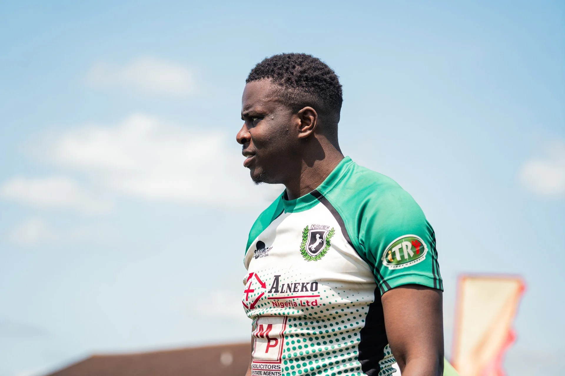 A male rugby player in a green and white jersey with sponsor logos, standing outdoors against a blue sky with clouds.