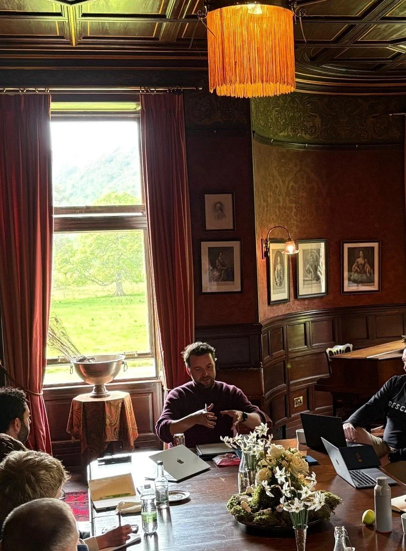 A group of people having a meeting in a room with large window, red curtains, and ornate wooden walls. A man at the head of the table is gesturing as he speaks. The table has laptops, water bottles, flowers, and notebooks.