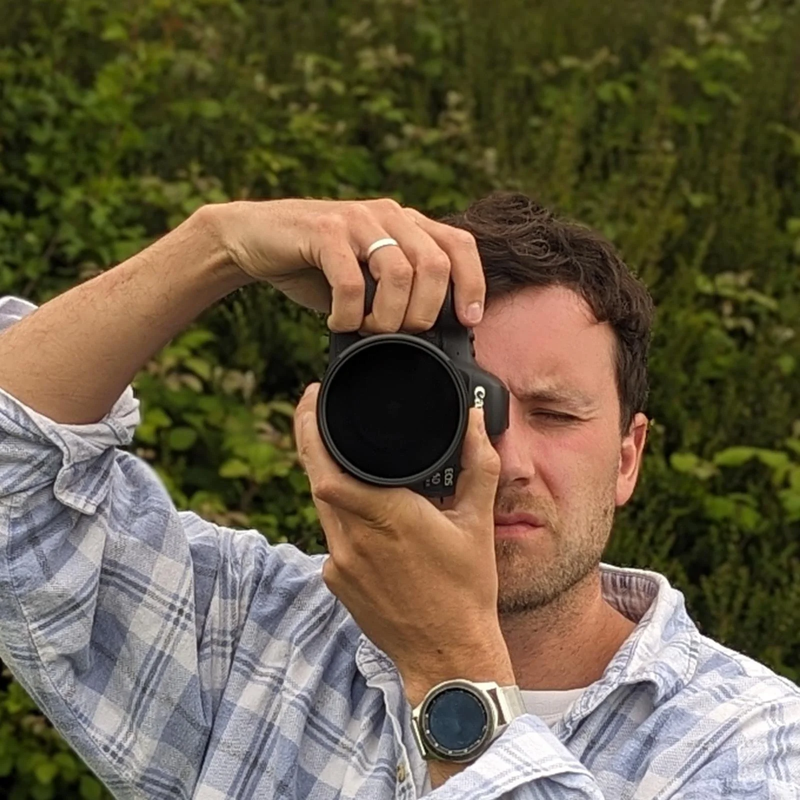A man is taking a photo with a camera outdoors, with greenery in the background, wearing a light-colored plaid shirt and a watch on his left wrist.