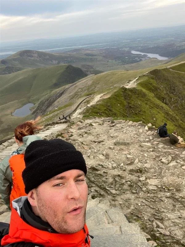 Man taking a selfie on a mountain trail with hikers in the background, overlooking green hills and a distant body of water under a cloudy sky.