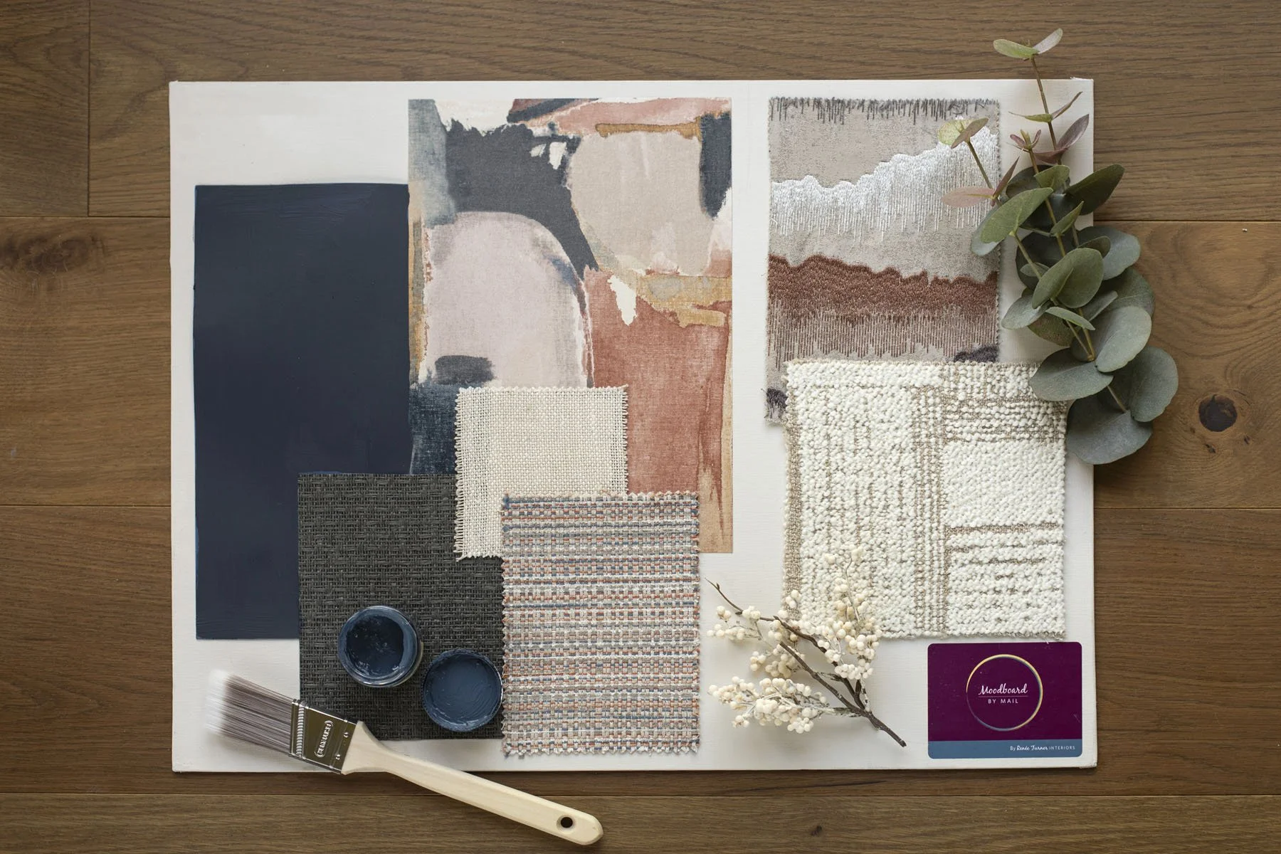 A flat lay of fabric swatches, paint samples, and decorative elements on a wooden surface, including a paintbrush, small paint containers, fabric samples, and sprigs of greenery and white berries.