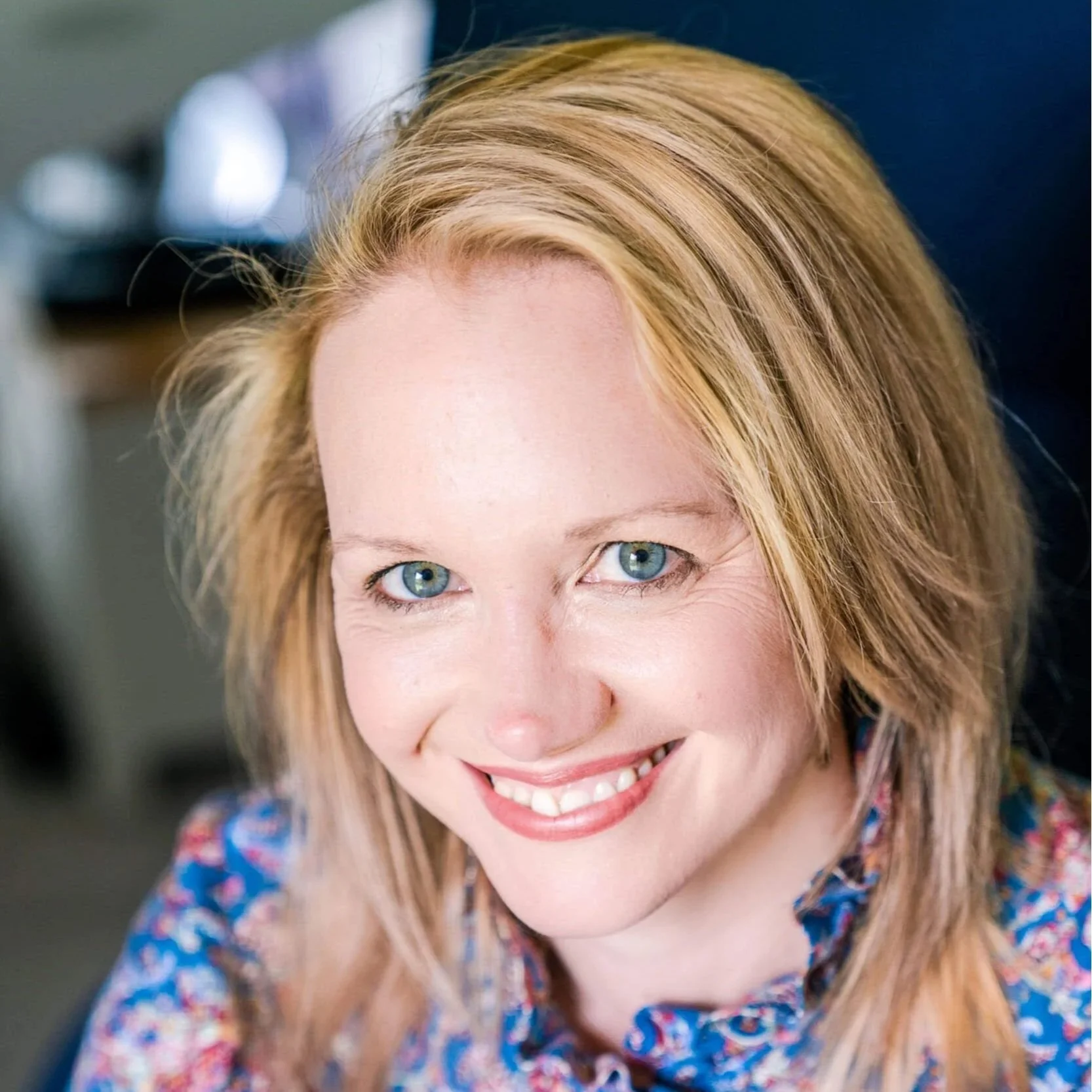 Close-up of a smiling woman with blonde hair and blue eyes, wearing a colorful patterned top.