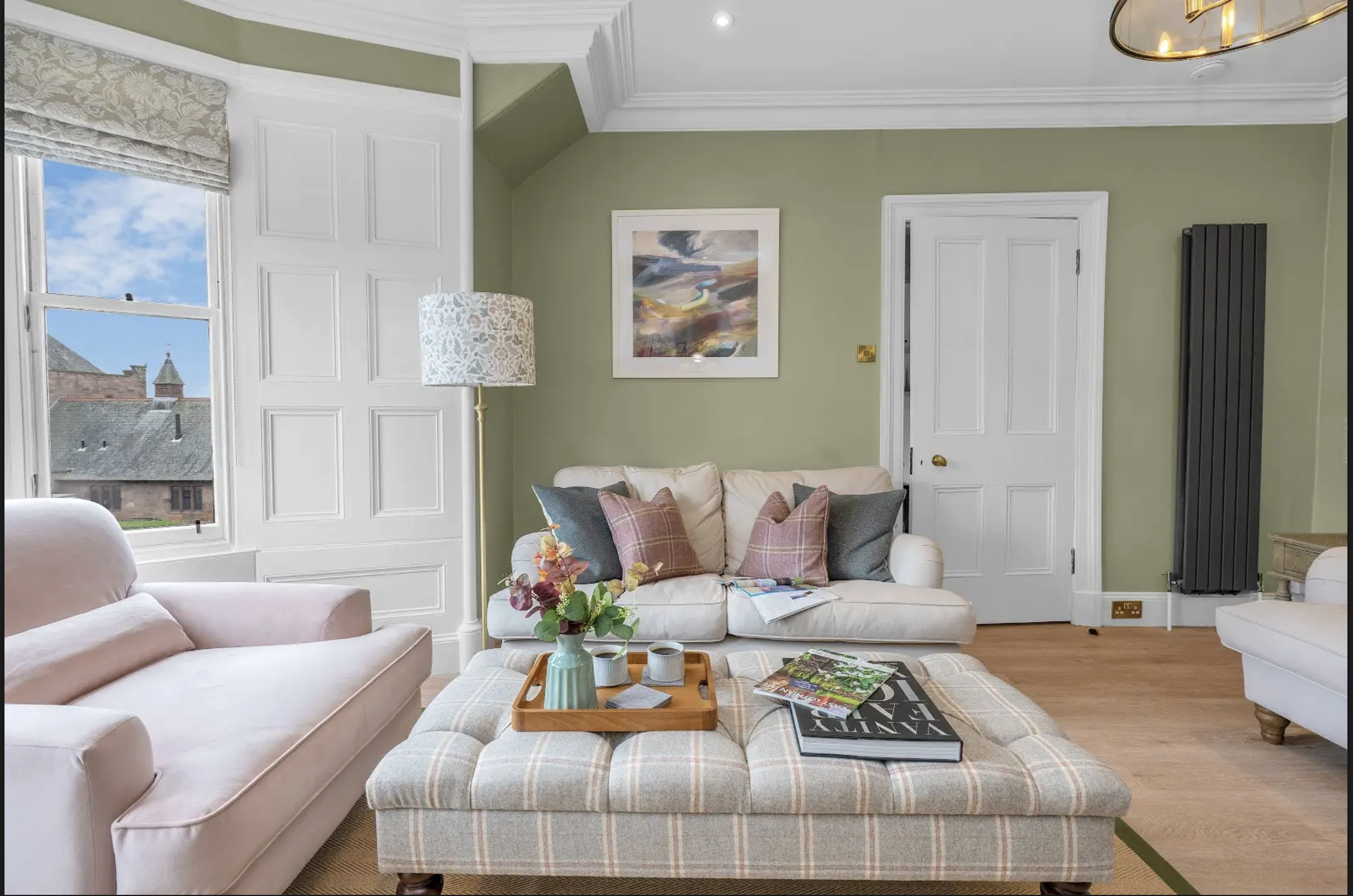 Bright living room with large window, teal armchair, gray sofa with colorful cushions, green knitted pouf, and indoor plants on windowsill and wall shelves.