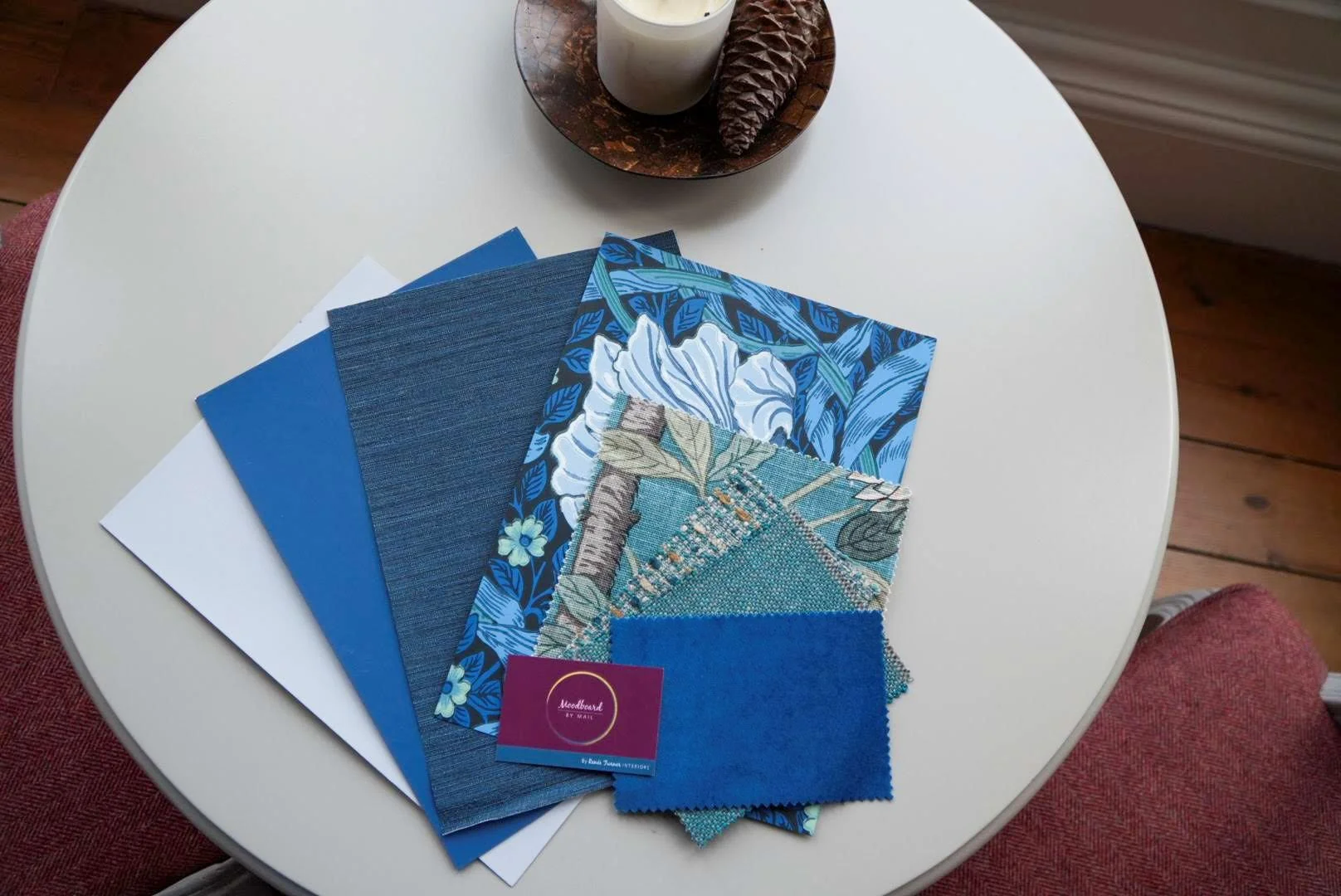 Swatches of blue and patterned fabric, a business card, and textile samples on a white circular table, with a candle and pine cone on a wooden tray in the background.