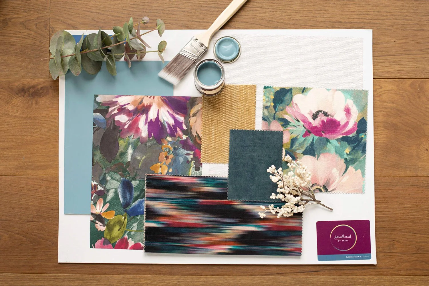 A flat lay of interior design samples including fabric swatches with floral patterns, paint samples, a small branch with white berries, a can of blue paint, a paintbrush, and a mood board card on a wooden surface.