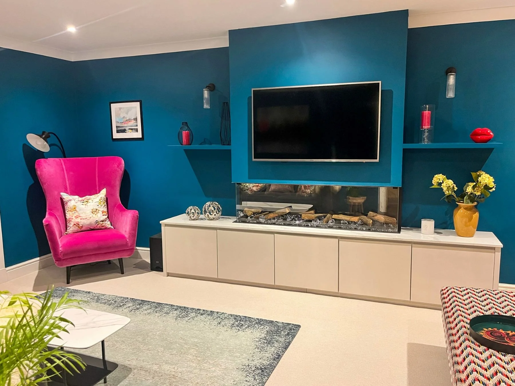 Modern living room with teal walls, pink velvet armchair with floral cushion, white cabinet with glass fireplace, large flat-screen TV, wall-mounted shelves with red and yellow decor, yellow vase with flowers, gray and white rug, and patterned ottoma