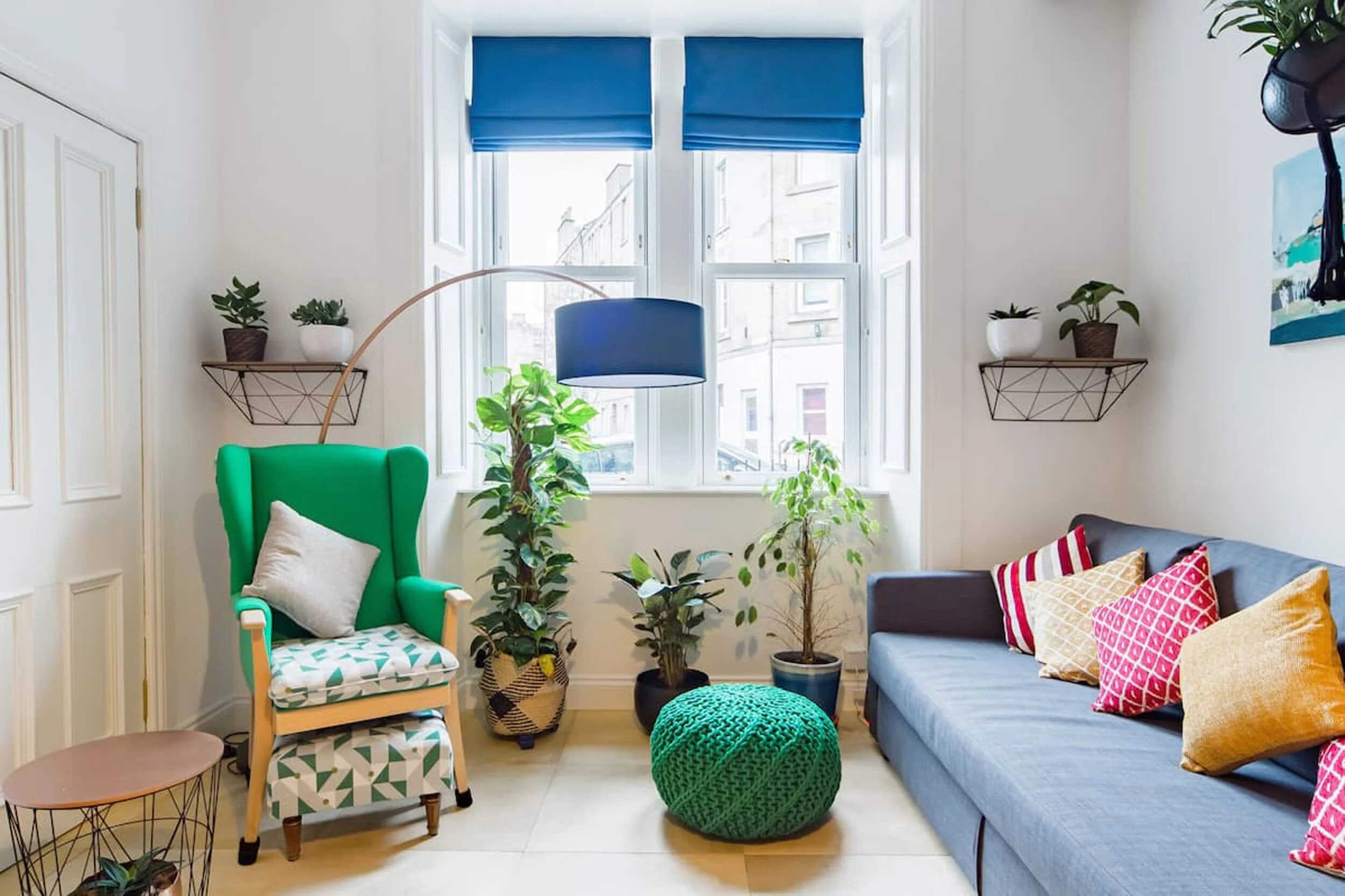 Bright living room with a gray sofa adorned with colorful pillows, a green armchair with a white pillow, potted plants, a round green pouf, a window with blue shades, and wall shelves with potted plants.