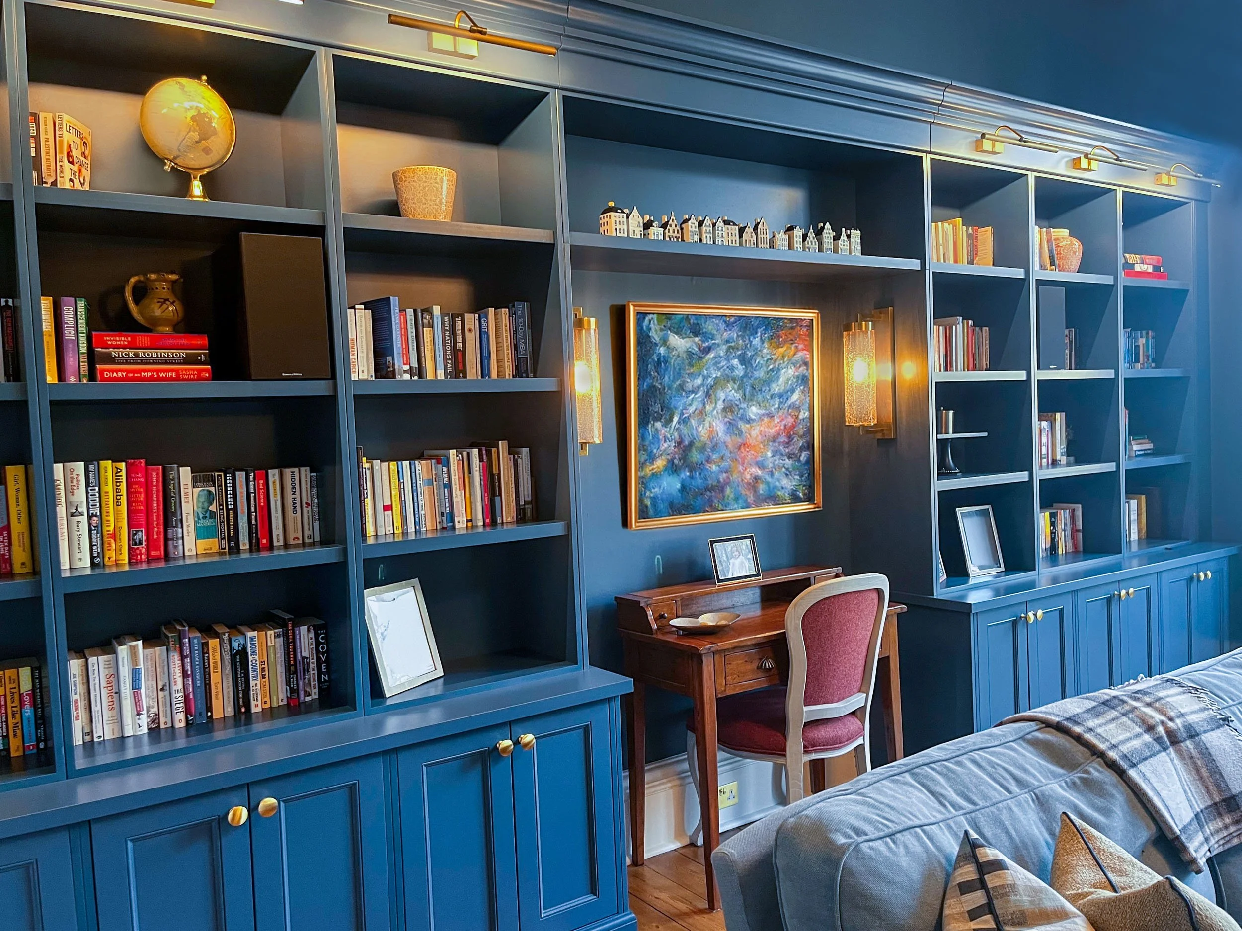 A living room with a blue bookshelf wall, filled with books, a globe, and decorative items. There's a small wooden desk with a red upholstered chair, a framed abstract painting, and a sofa with pillows in front.