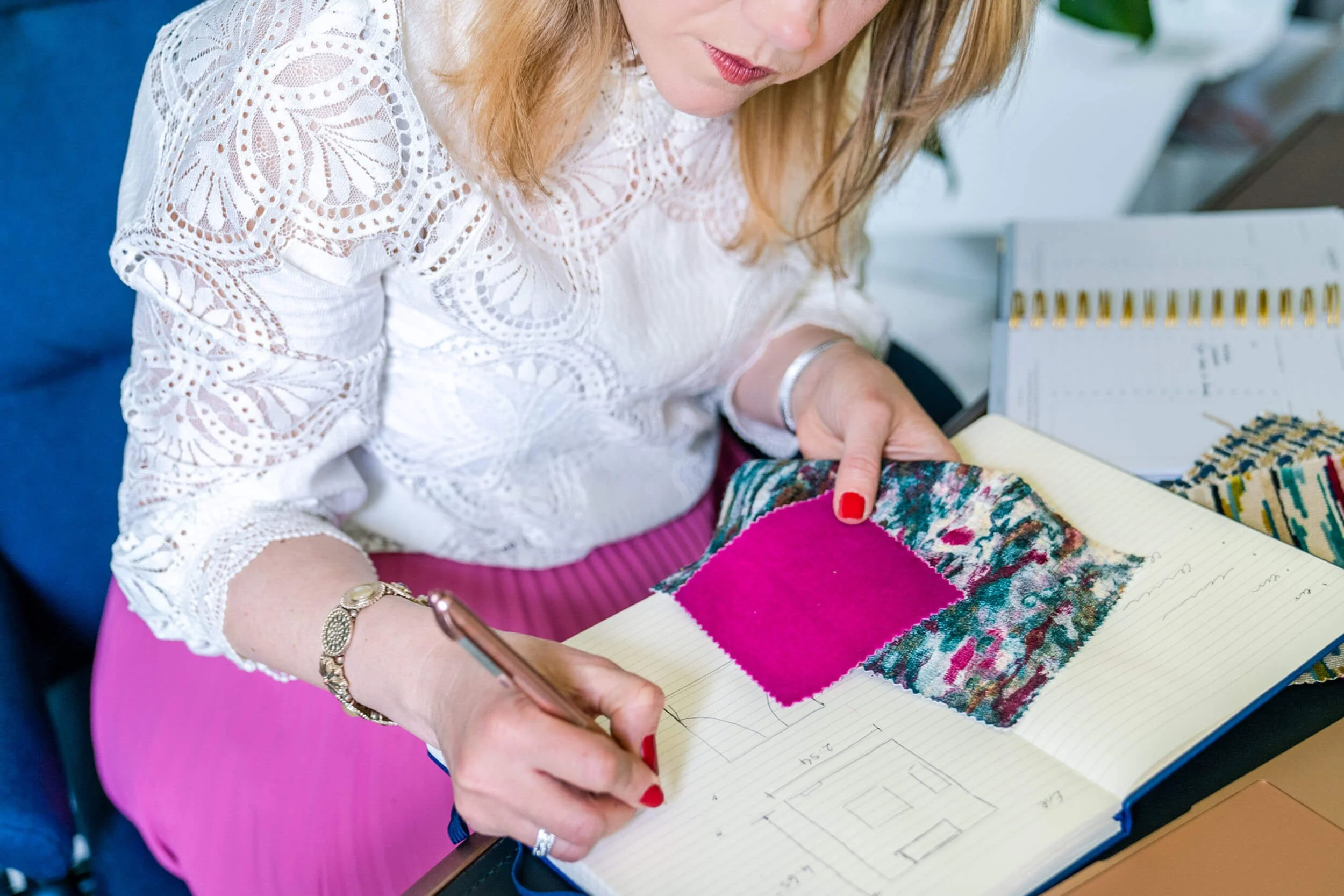 A woman with red hair, wearing a white lace blouse and pink skirt, is sitting at a desk reviewing fabric samples and sketching notes in a notebook, with additional fabric swatches and a calendar in the background.