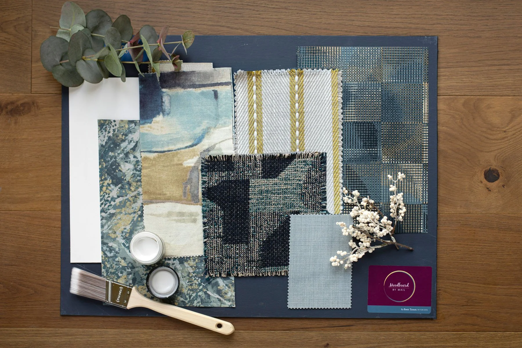 Color palette and fabric swatches for interior design, with flowers, a brush, and a Moodboard card on a wooden surface.