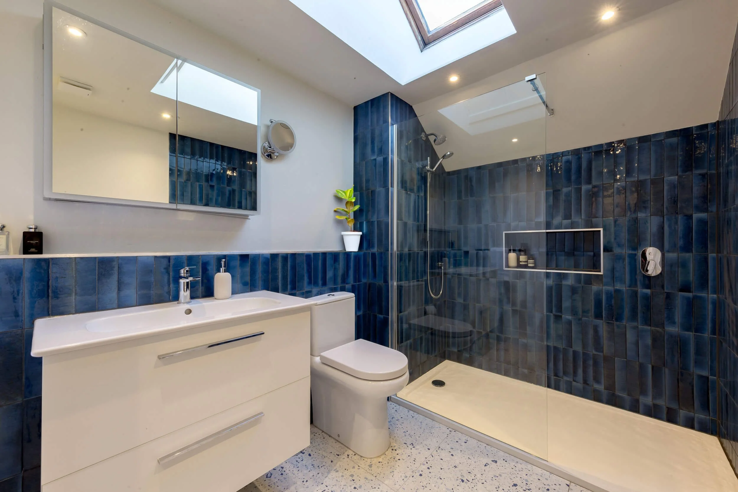 Modern bathroom with blue tiled walk-in shower, white vanity with sink, toilet, large mirror, skylight, and decorative plant.