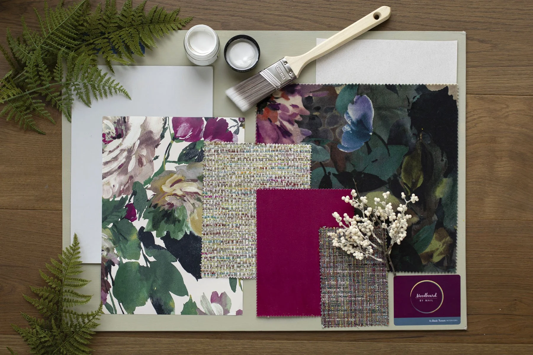 Painting or fabric samples, paintbrush, paints, fabric swatches, and greenery on a wooden surface, likely in a design or decorating setting.