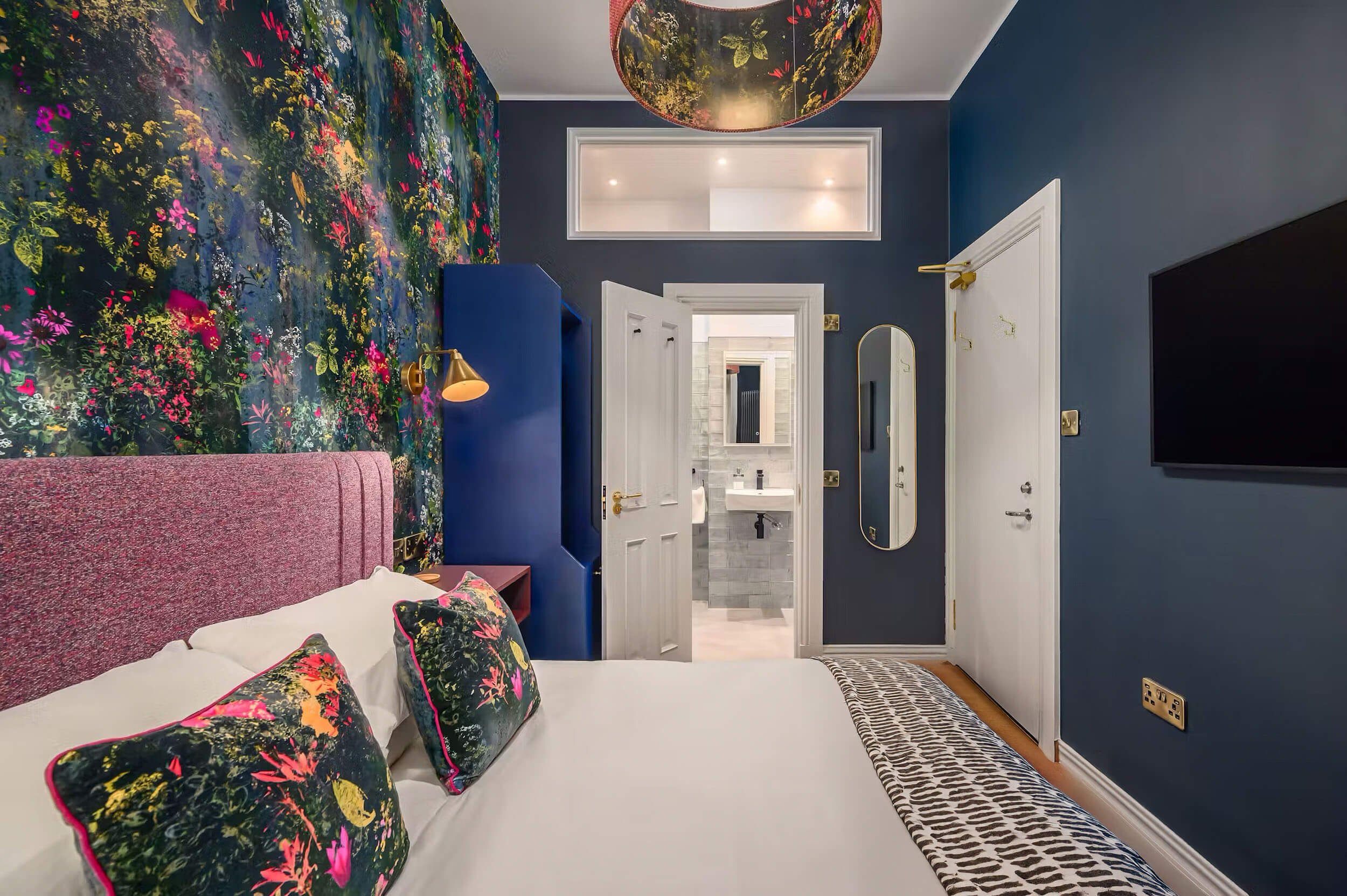 A colorful bedroom with a floral wallpaper accent wall, a pink upholstered bed with matching pillows, dark blue walls, a wall-mounted gold reading lamp, a white door leading to an en suite bathroom, a large mirror, and a black flat-screen TV.