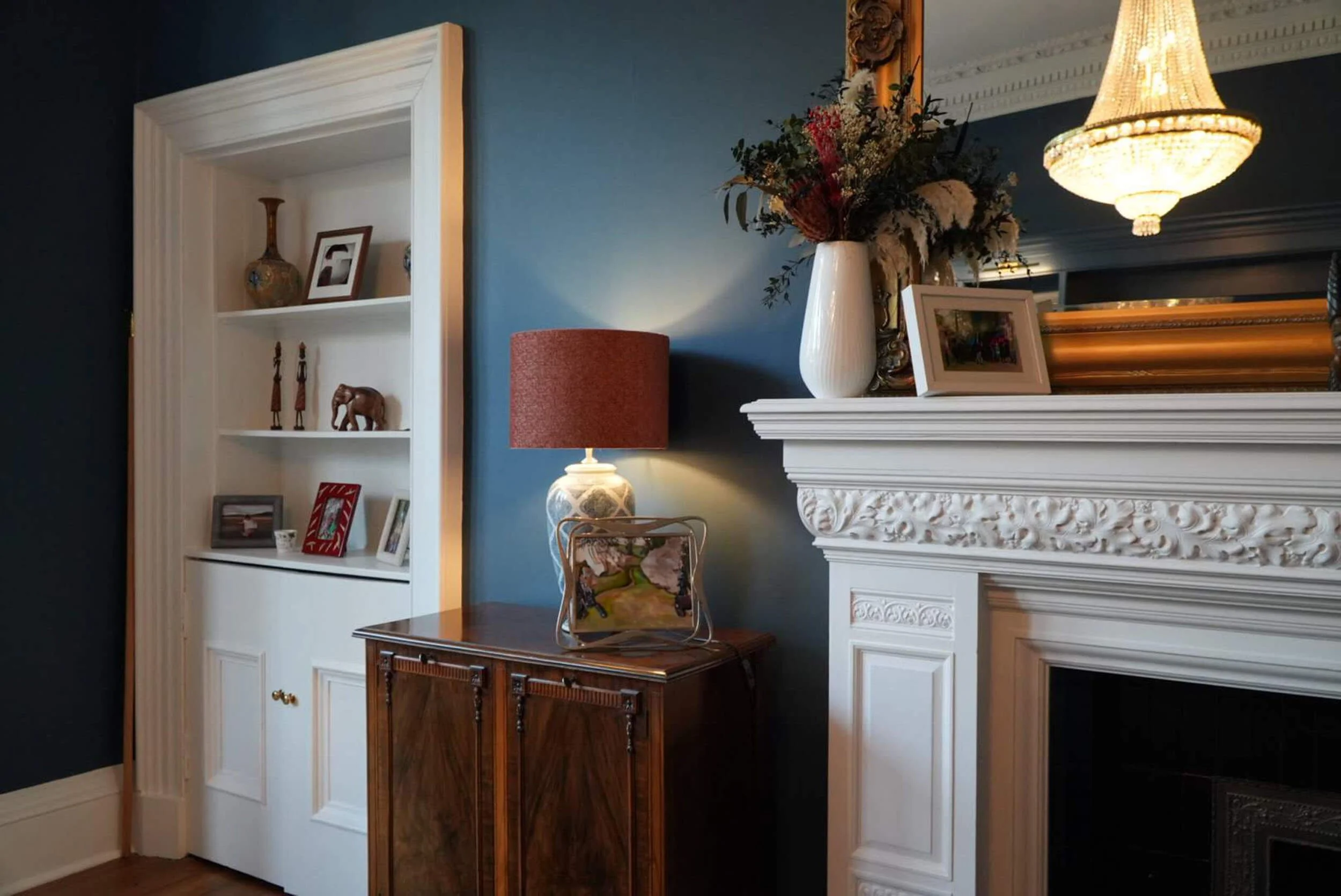 Living room interior with a white built-in bookshelf, a wooden cabinet with a table lamp, a white vase with flowers, a framed photo, and a decorative mirror above a white fireplace mantel.
