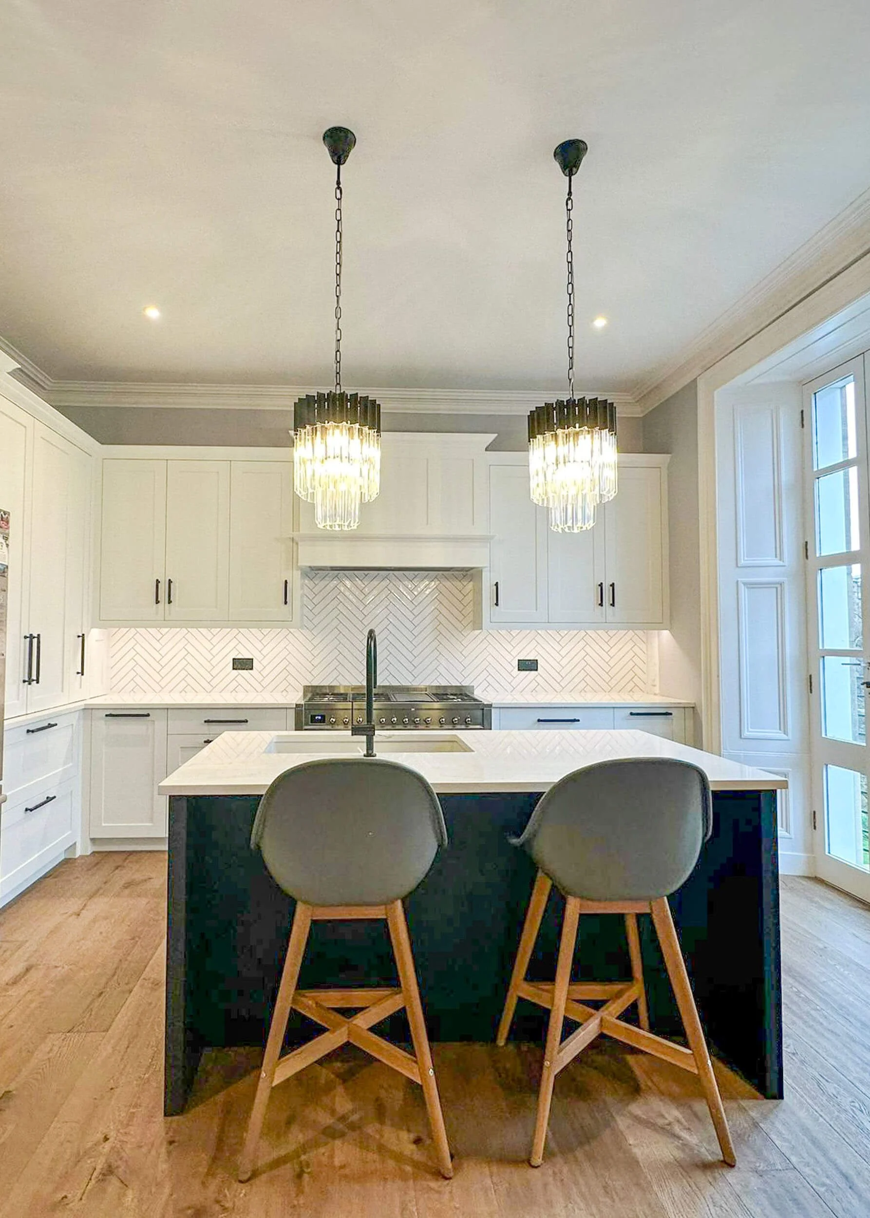 Modern kitchen with two hanging pendant lights, white cabinets, a white herringbone tile backsplash, a central island with a sink, two gray chairs with wooden legs, and large windows.