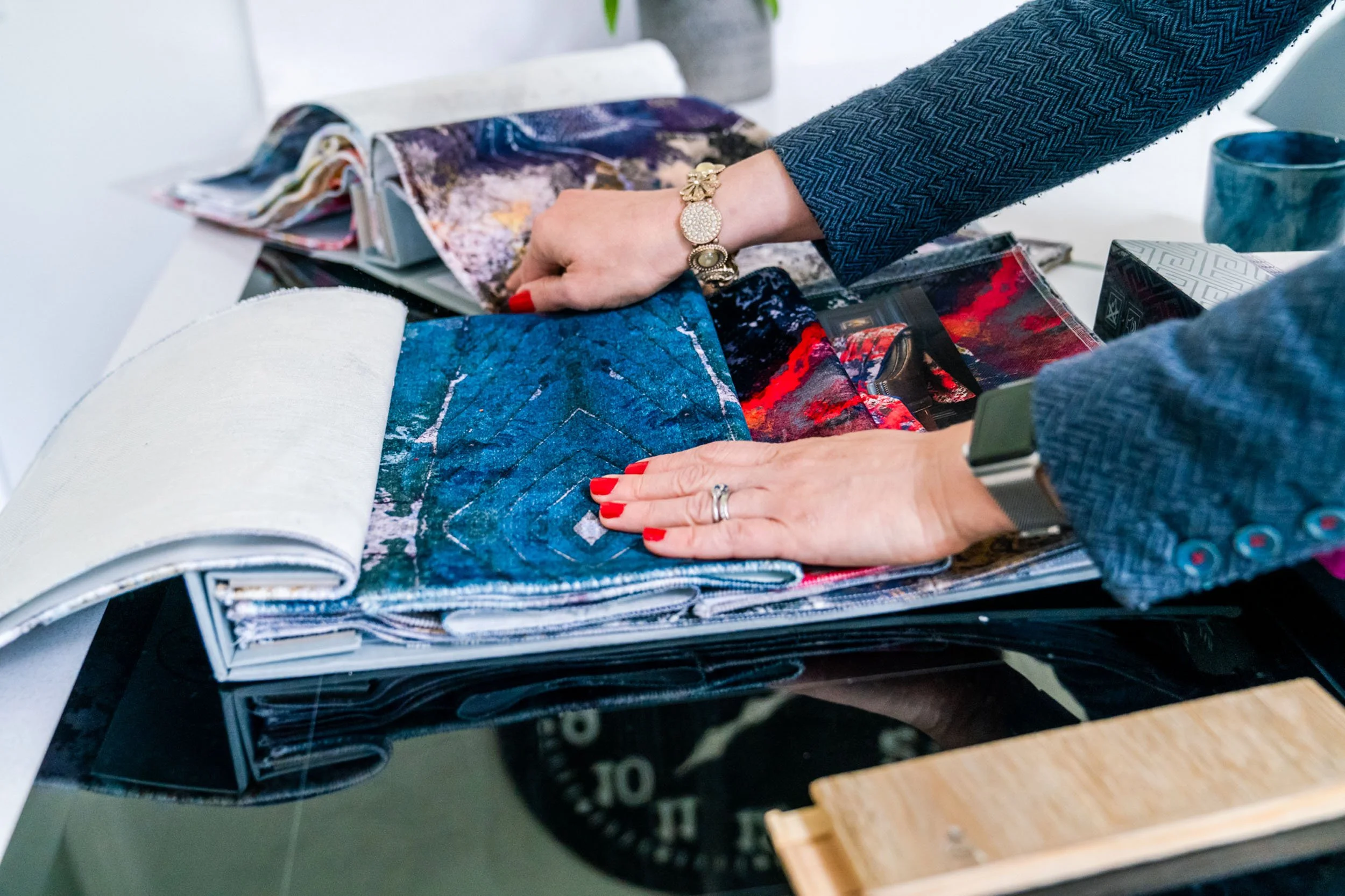 Person examining fabric samples and swatches on a table, with an open fabric catalog, wearing jewelry and a smartwatch.