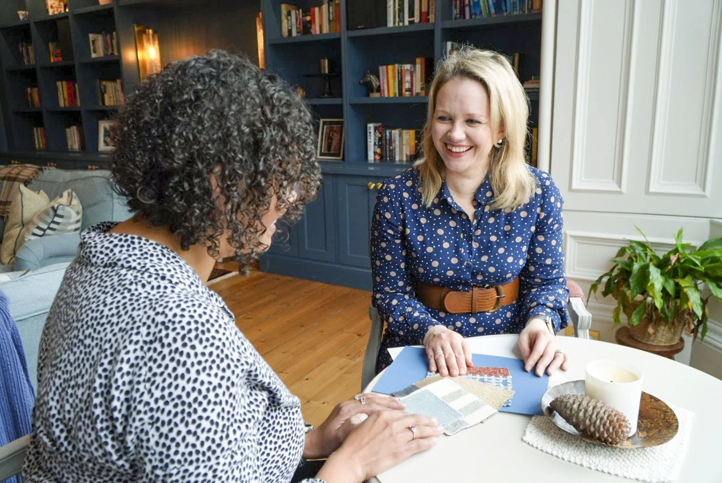 Two women sitting at a table, discussing fabric samples and design ideas in a cozy, well-decorated room with bookshelves, a blue cabinet, and a potted plant.