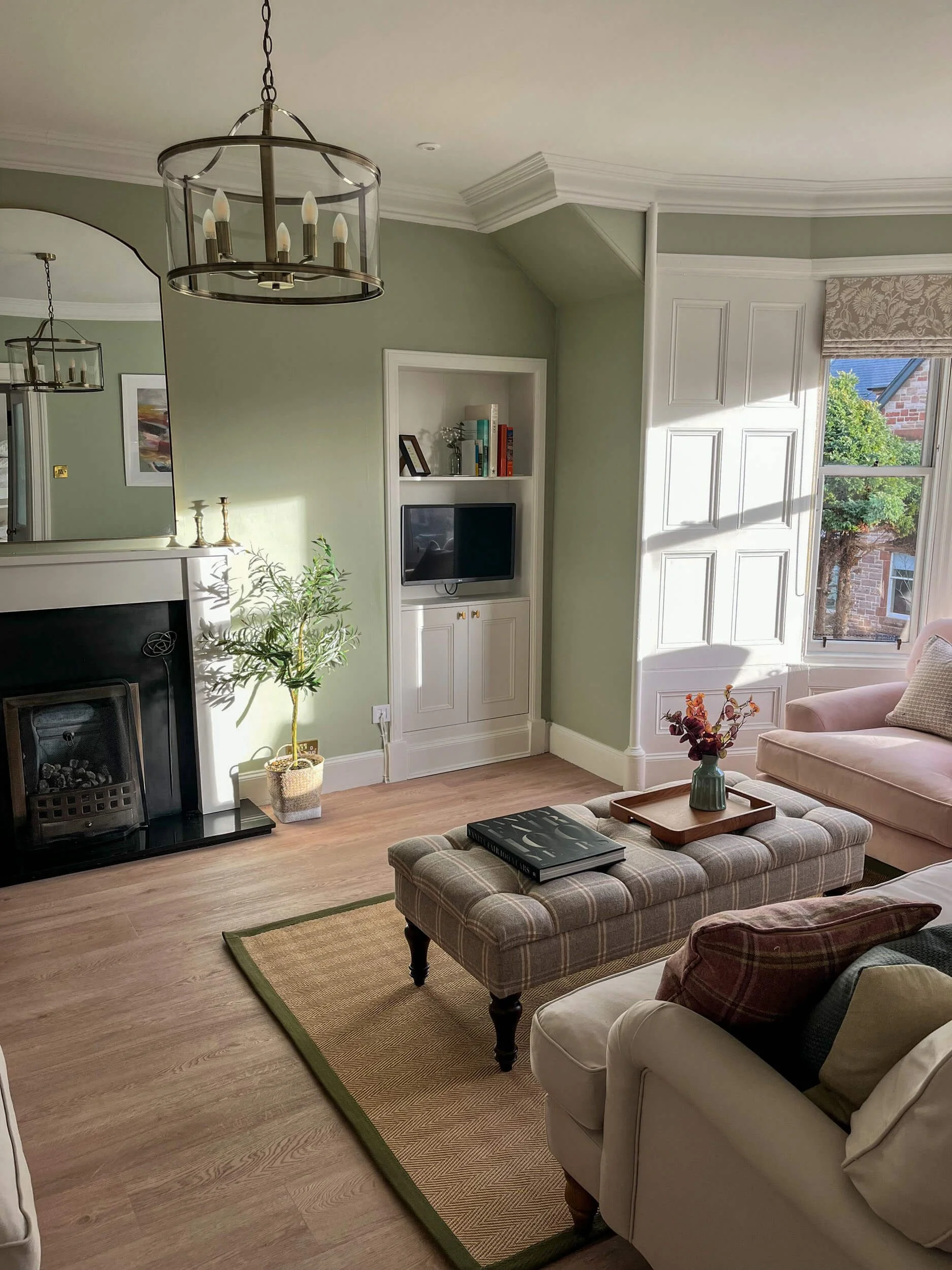 Living room with a fireplace, mirror, bookshelf, small TV, plant, chandelier, window, and furniture including a beige couch, plaid ottoman with books, and a pink armchair.