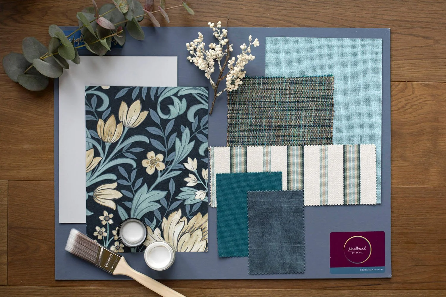 Color palette and fabric samples for interior design placement, including blue and beige floral wallpaper, fabric swatches, paint, and accessory samples, on a blue board on a wooden surface.