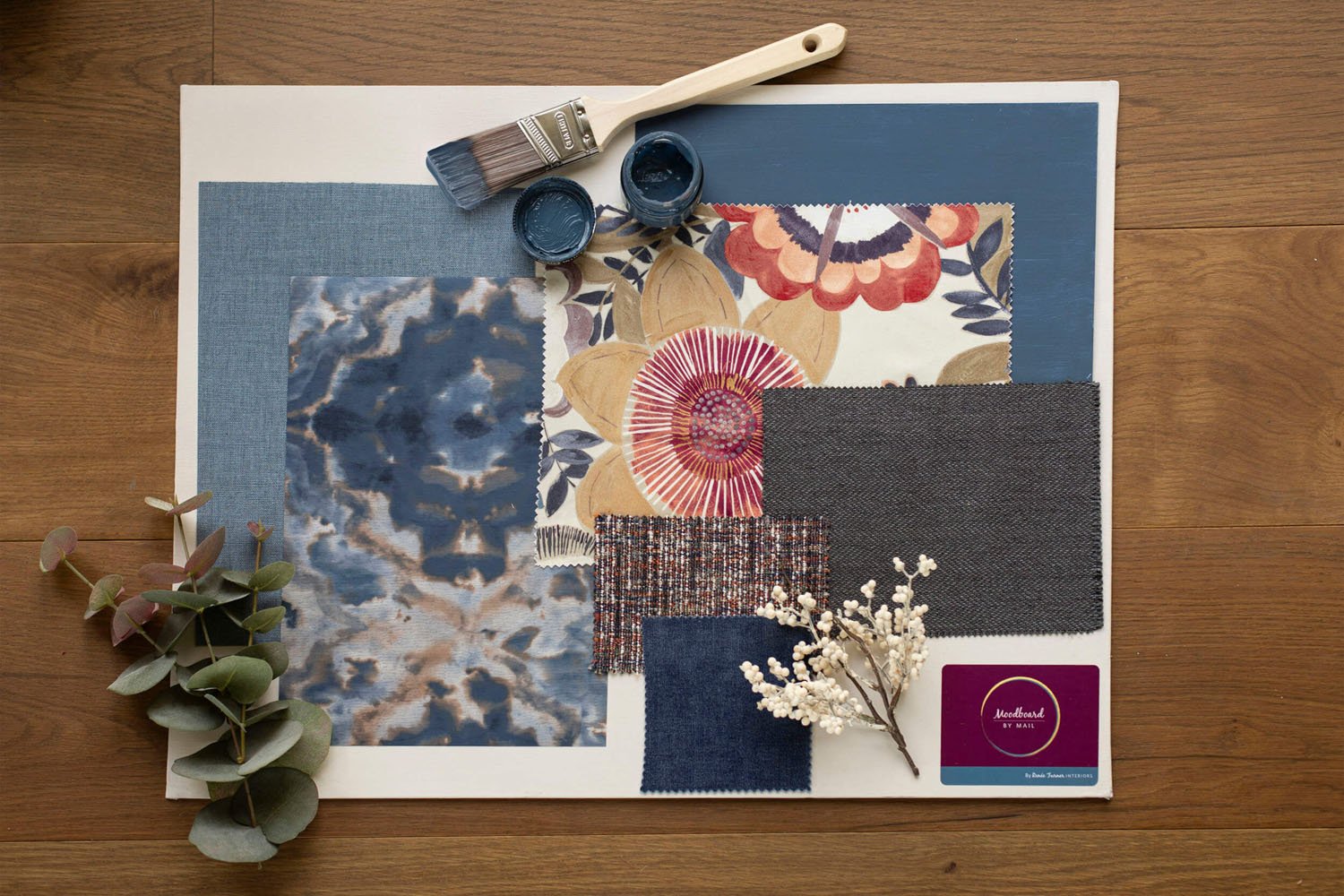 A flat lay of fabric swatches, paint samples, and decorative elements on a wooden surface, including a paintbrush, small paint containers, fabric samples, and sprigs of greenery and white berries.