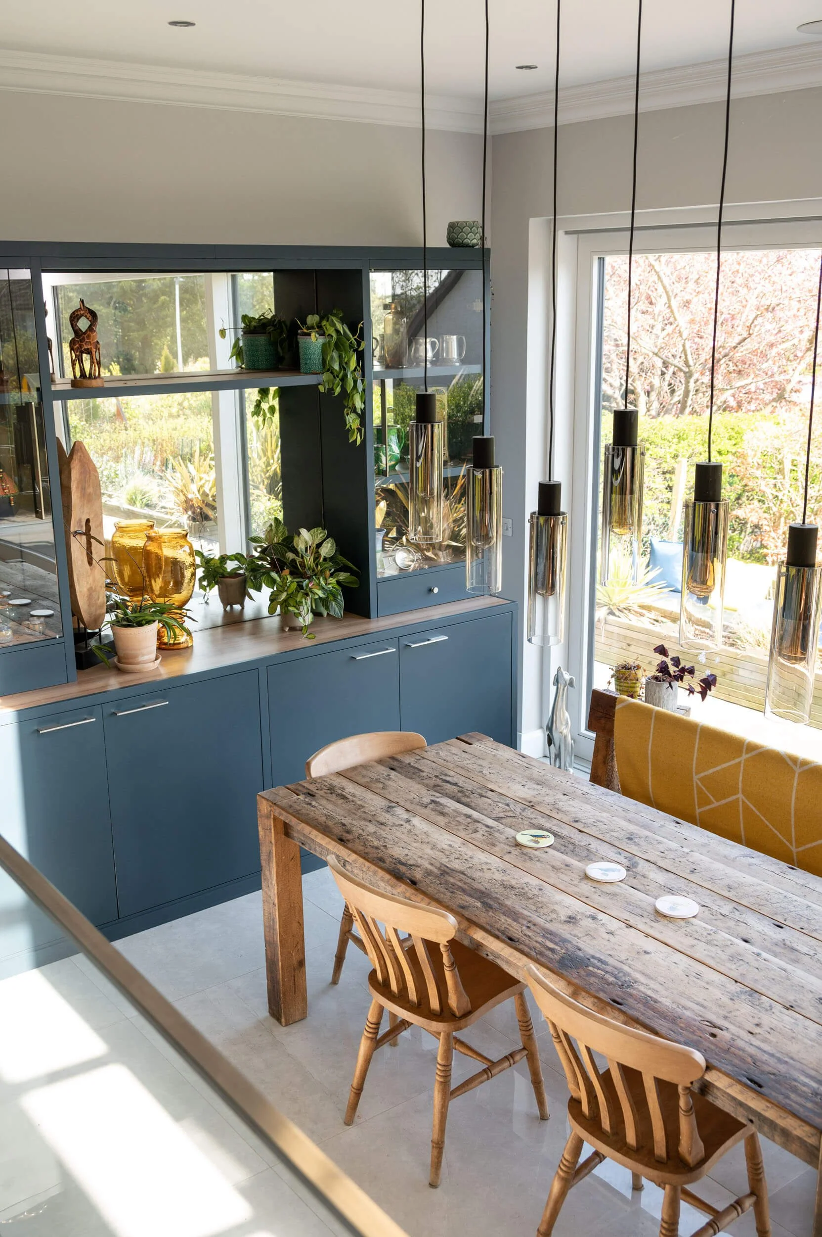 A dining area with a rustic wooden table, three wooden chairs, blue cabinetry with plants and decorative vases, hanging pendant lights, and large windows showing a garden outside.