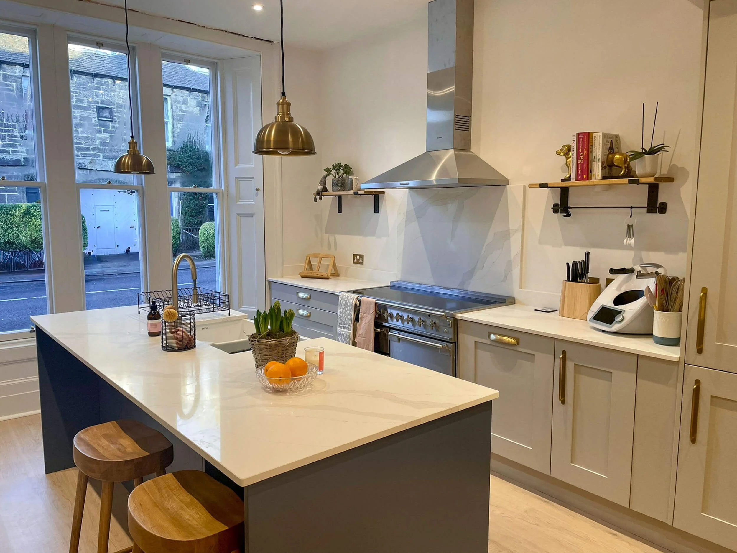 Modern kitchen with white cabinets, marble countertops, a kitchen island with two wooden stools, and large windows showing an outdoor street view. The kitchen has a stainless steel oven and range, a brass pendant light, and decorative shelves with books and plants.