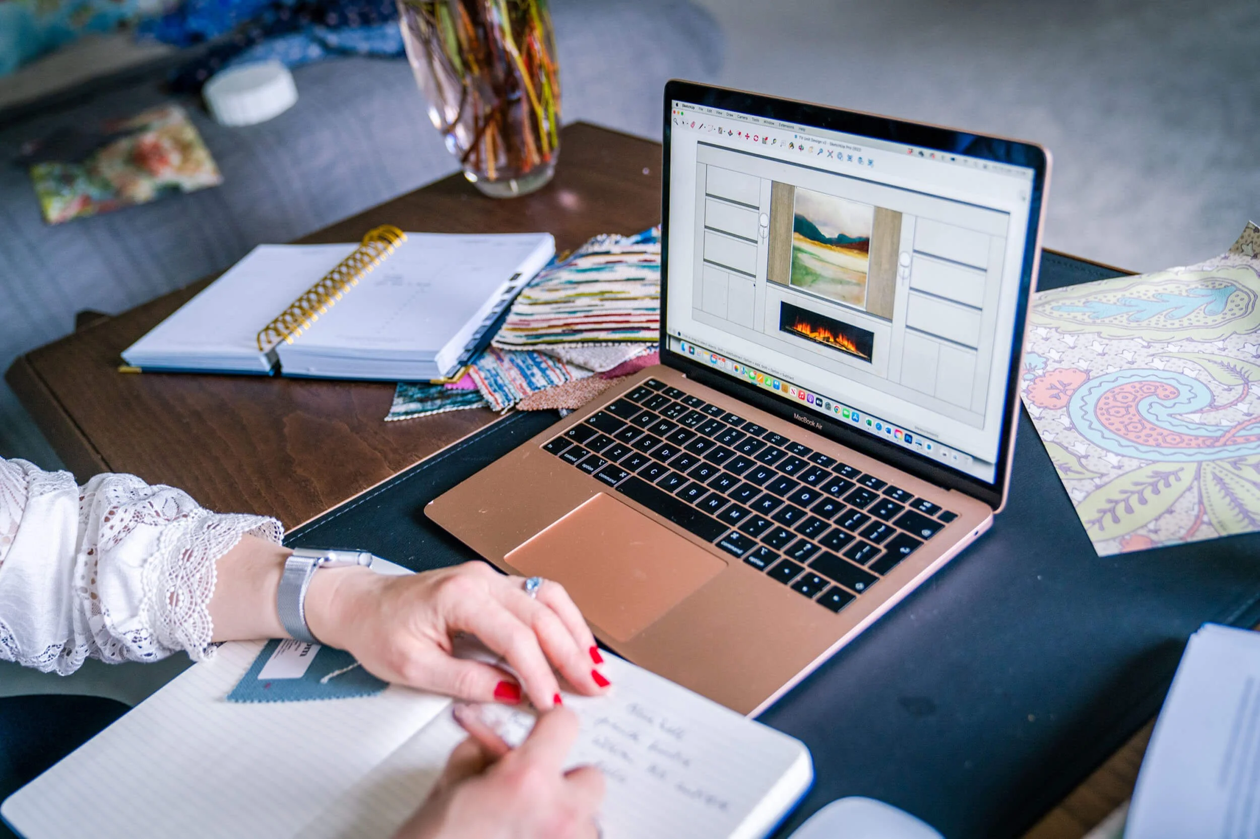 A person's hand with red fingernail polish takes notes near a closed laptop on a dark desk. The laptop screen displays a design program. There are open notebooks, a colorful woven fabric, a potted plant, and various papers on the desk.