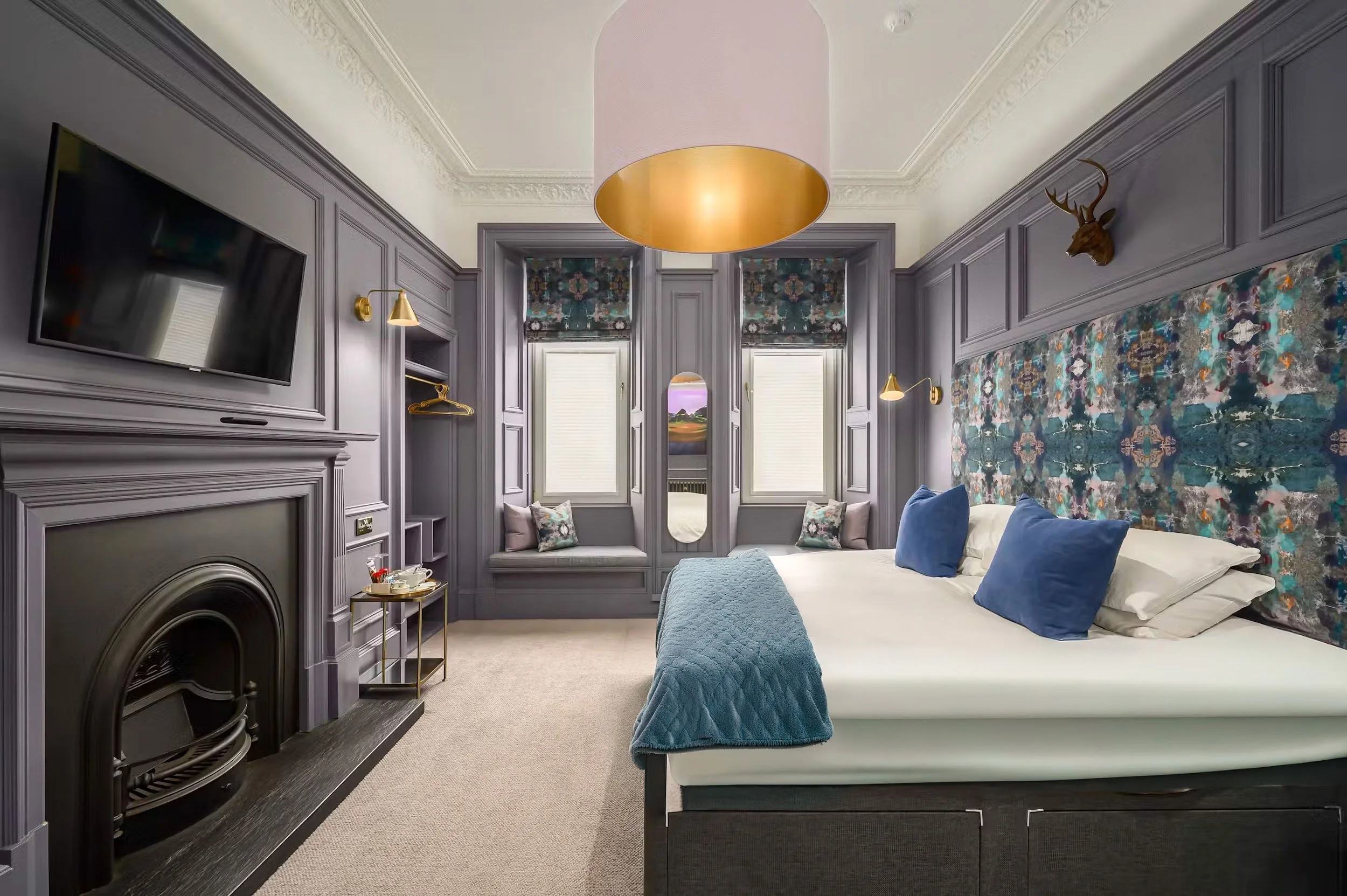 A bedroom with purple paneled walls, a large bed with blue pillows and a quilt, built-in window seats, a fireplace, a wall-mounted TV, and decorative elements like a deer head, gold wall lamps, and floral window coverings.