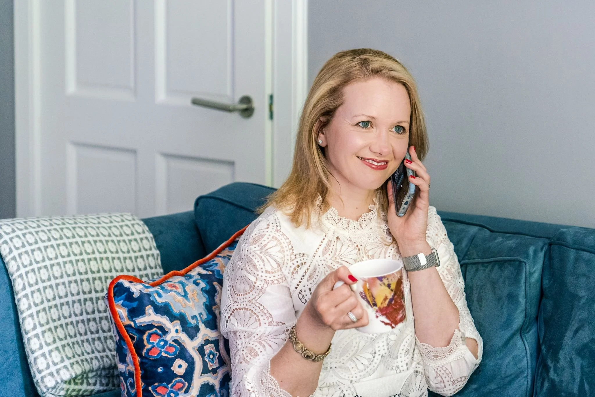 A woman with shoulder-length blonde hair, sitting on a blue couch, smiling and talking on her cell phone while holding a coffee mug in her other hand. She is wearing a white lace top, a watch, a bracelet, and earrings, with a patterned pillow behind her.