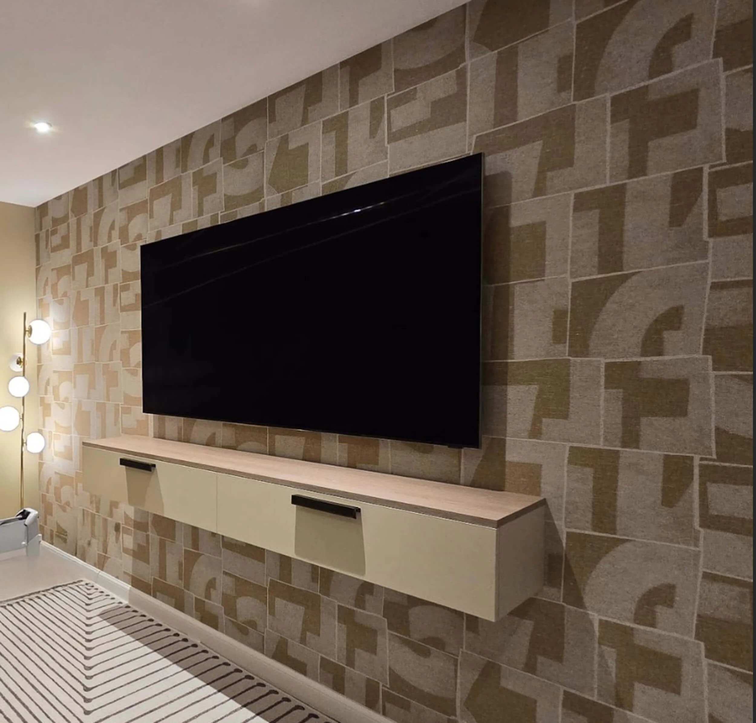 A modern living room with a large black flat-screen TV mounted on a patterned beige wall, and a floating light-colored wooden console beneath it.