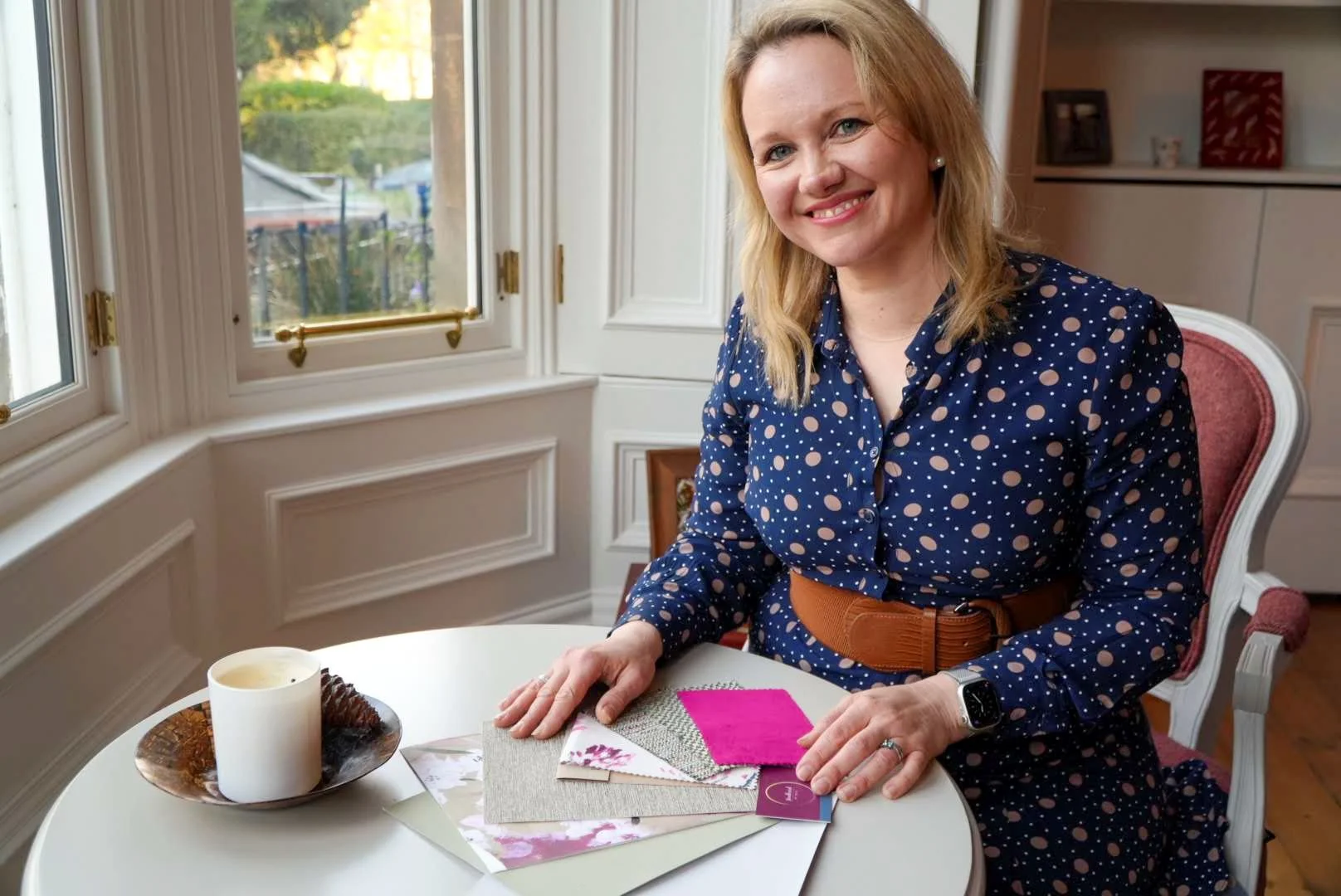 A woman with blonde hair, wearing a navy blue dress with white and beige polka dots, sitting at a round table in a room with large windows and white walls. The table has a candle and fabric swatches. She is smiling and looking at the camera.