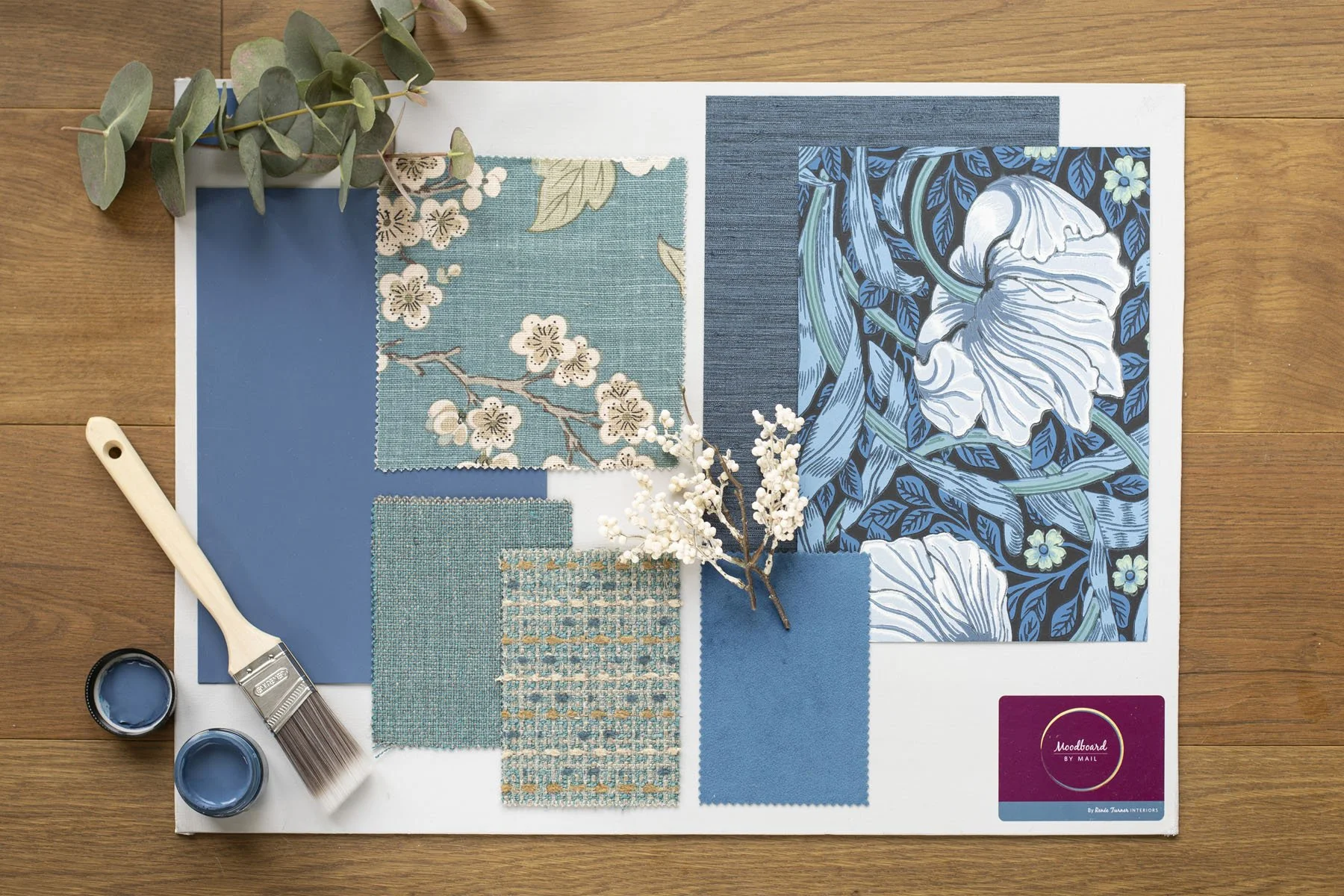 Color palette and fabric samples for interior design placement, including blue and beige floral wallpaper, fabric swatches, paint, and accessory samples, on a blue board on a wooden surface.