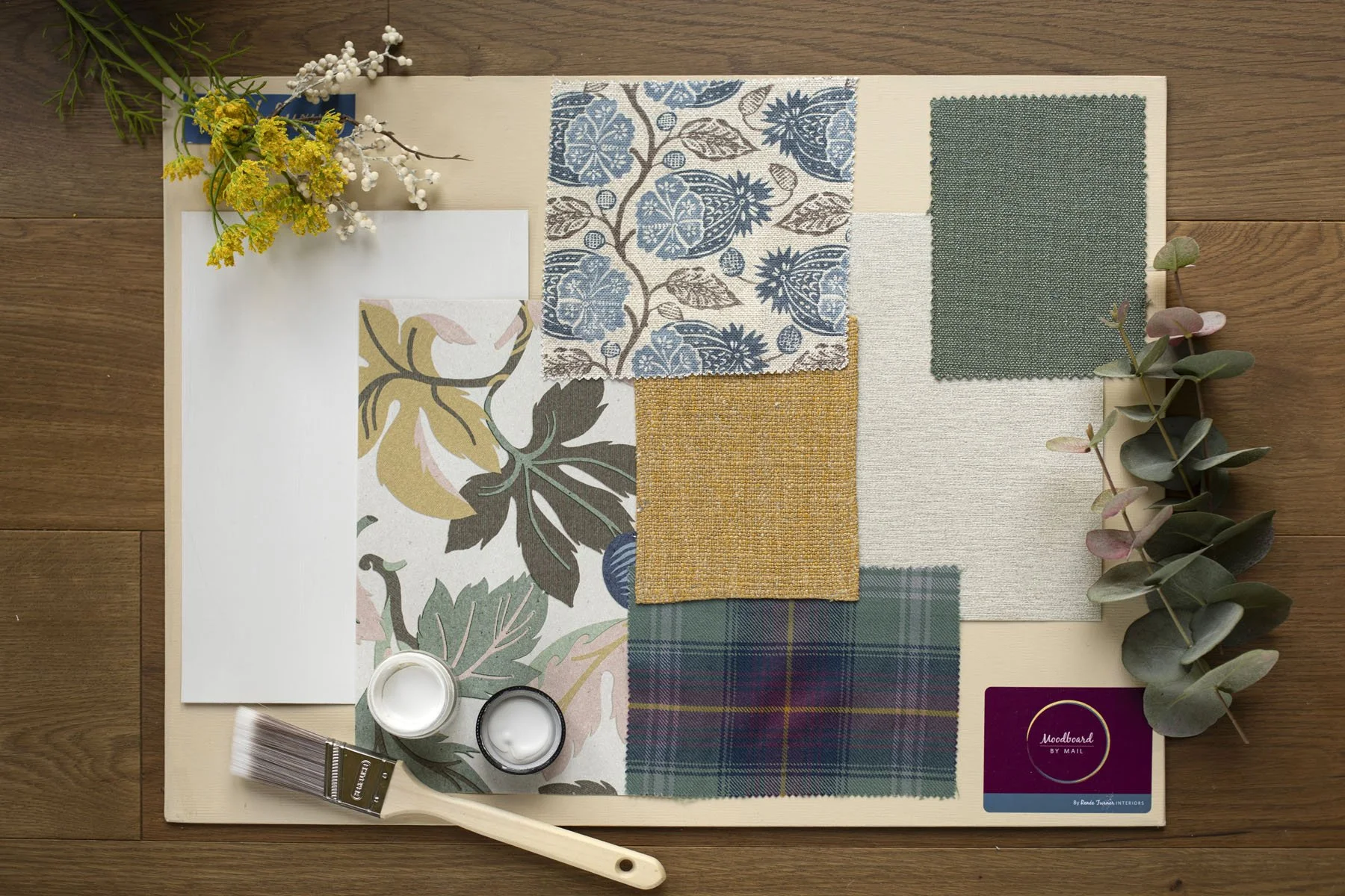 Interior design mood board with fabric swatches, floral cuttings, paint samples, and a colour palette on a wooden surface.