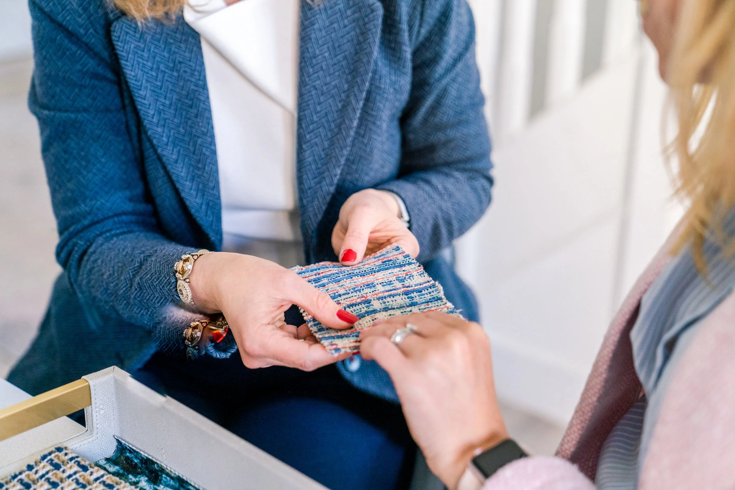 Two women are exchanging fabric samples, with one woman holding a colorful woven piece while the other is reaching to touch it, in a well-lit indoor setting.