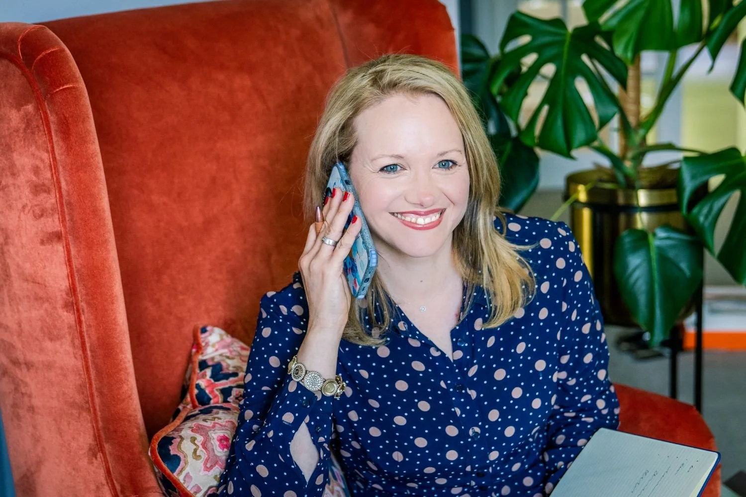 A woman with blonde hair and blue eyes smiling while talking on her cellphone, sitting on an orange velvet chair with a patterned pillow, in a room with large green houseplants.