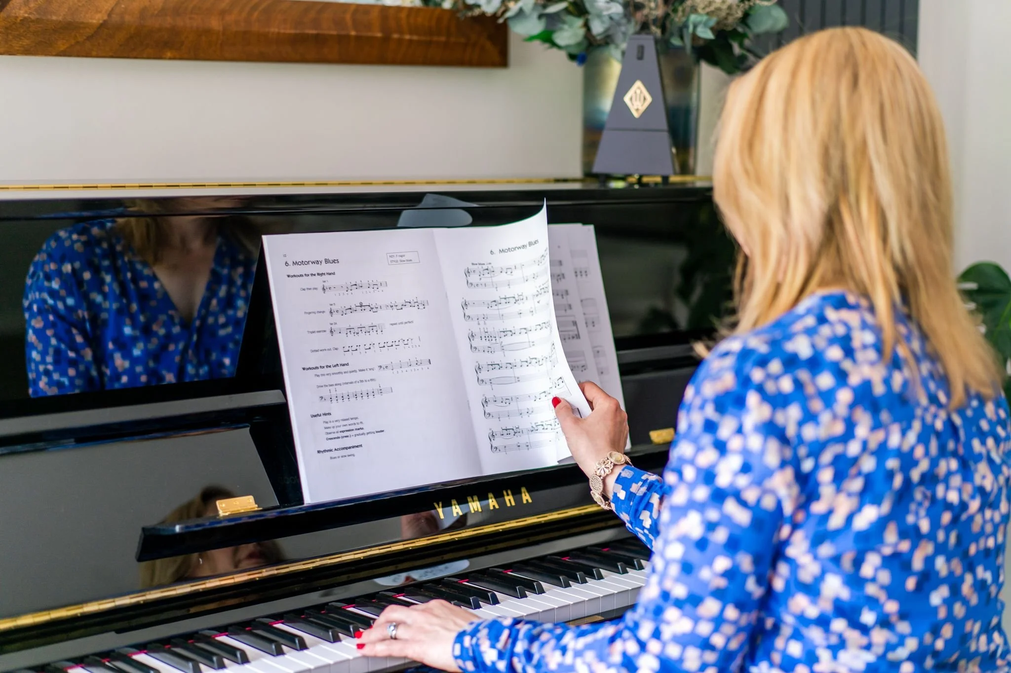 A woman with blonde hair playing a Yamaha black upright piano, reading sheet music titled 'Motorway Blues.' She is wearing a blue patterned shirt, a wristwatch, and a ring, with her nails painted red. In the background, there are plants and a decorative object on the piano.