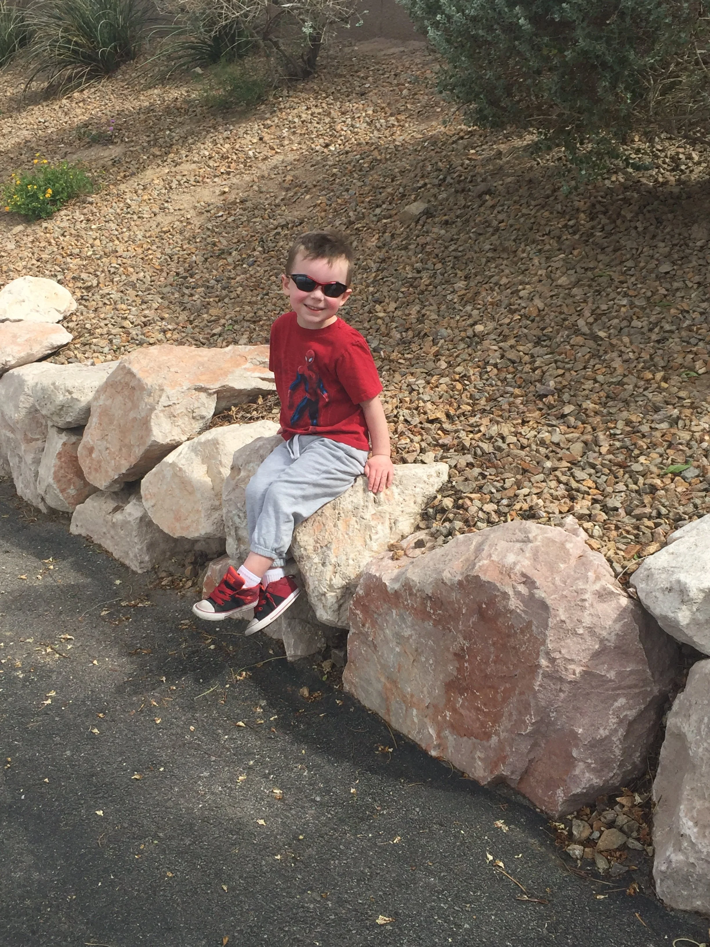Young child wearing sunglasses and a red shirt sitting on landscaping rocks beside a walking path.