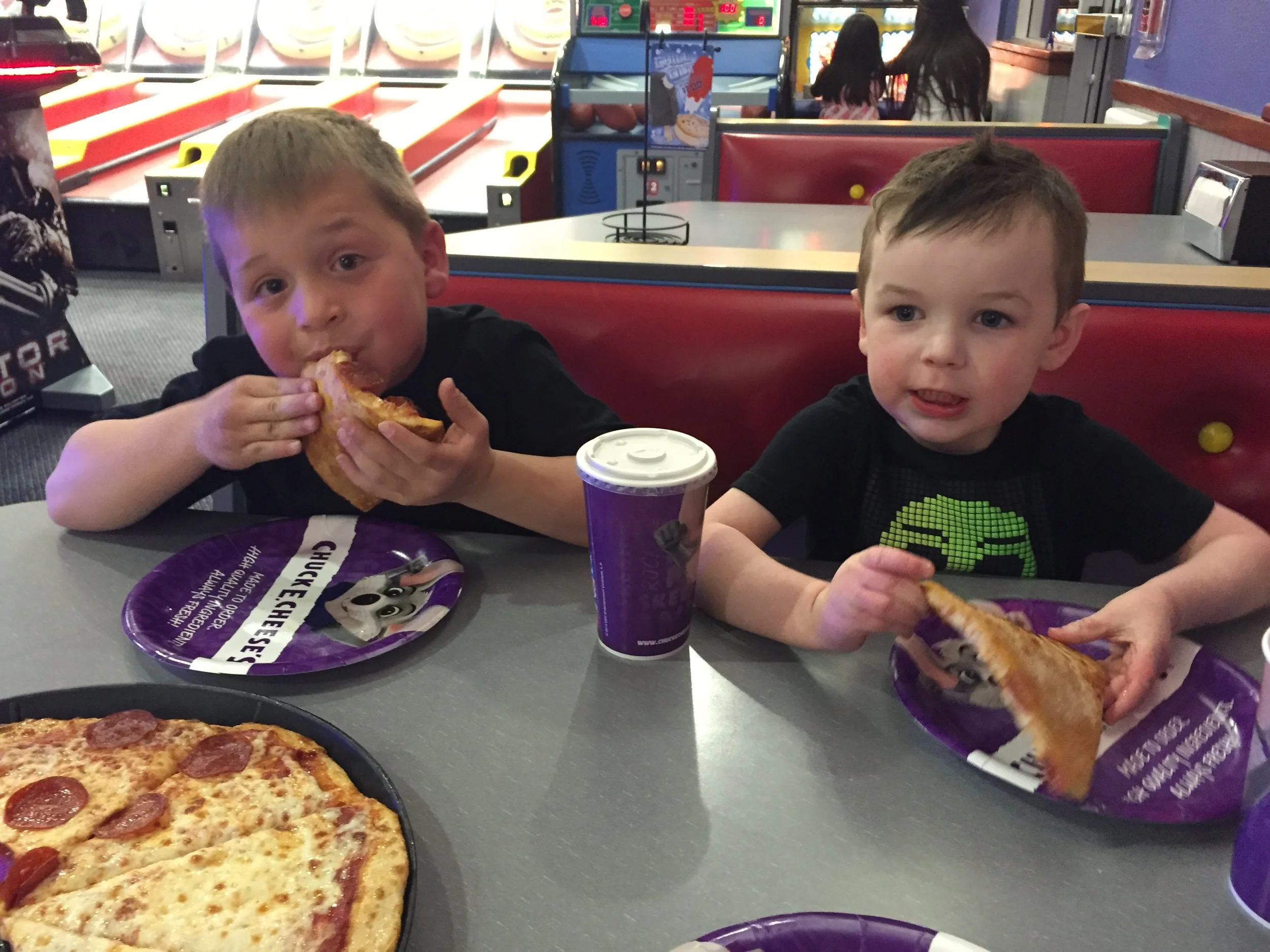 Two children seated at a Chuck E. Cheese table eating pizza, with arcade games visible in the background.