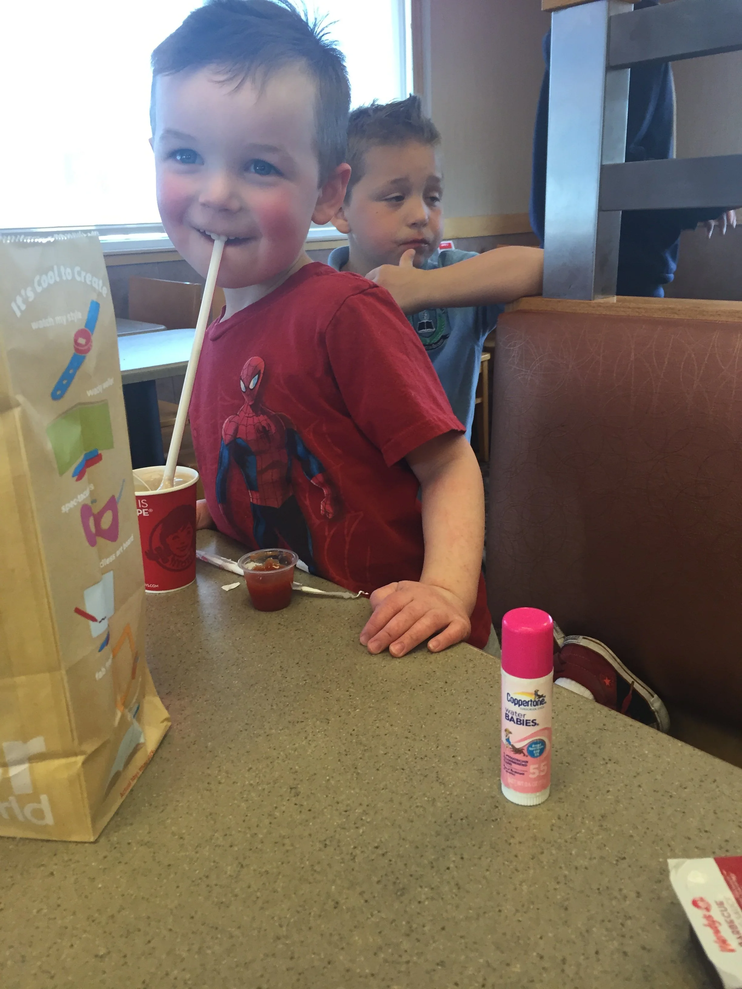 Young child in a red Spider-Man shirt at a restaurant table with an SPF 55 sunscreen stick visible in the foreground and another child seated behind him.