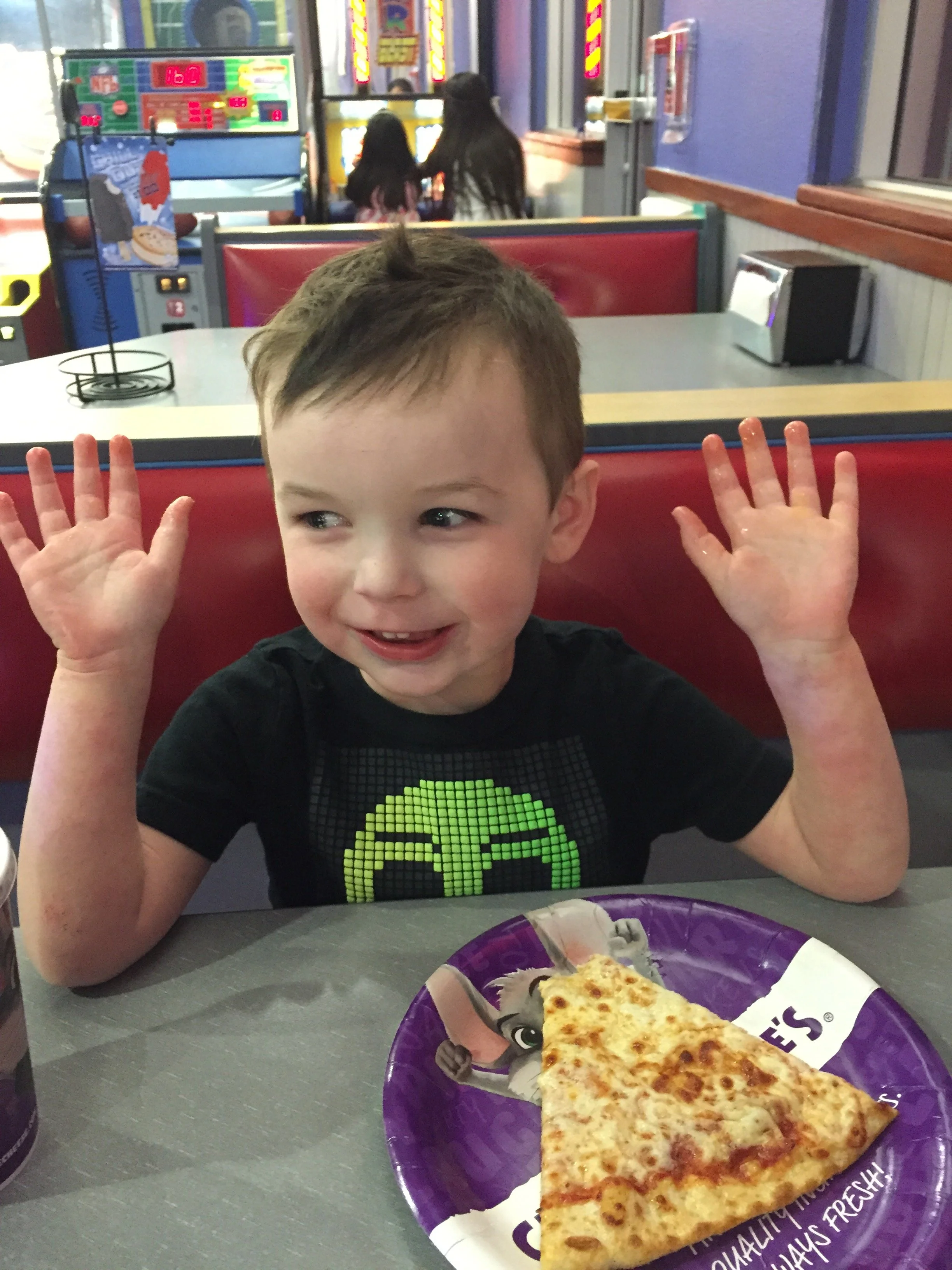 Young child smiling with both hands raised while seated at a Chuck E. Cheese table with a slice of pizza.