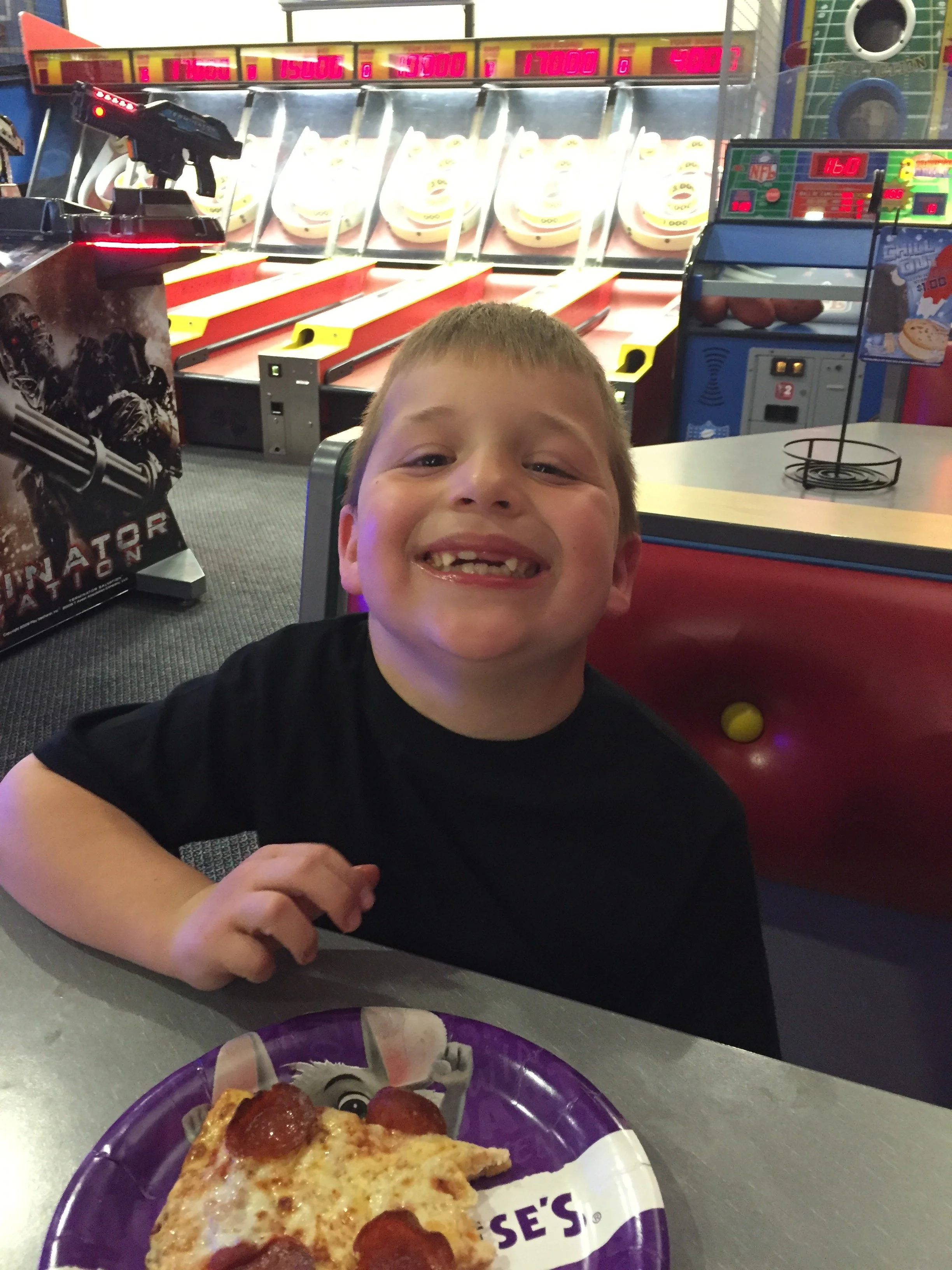 Older child smiling at a Chuck E. Cheese table with pizza and arcade games visible behind him.
