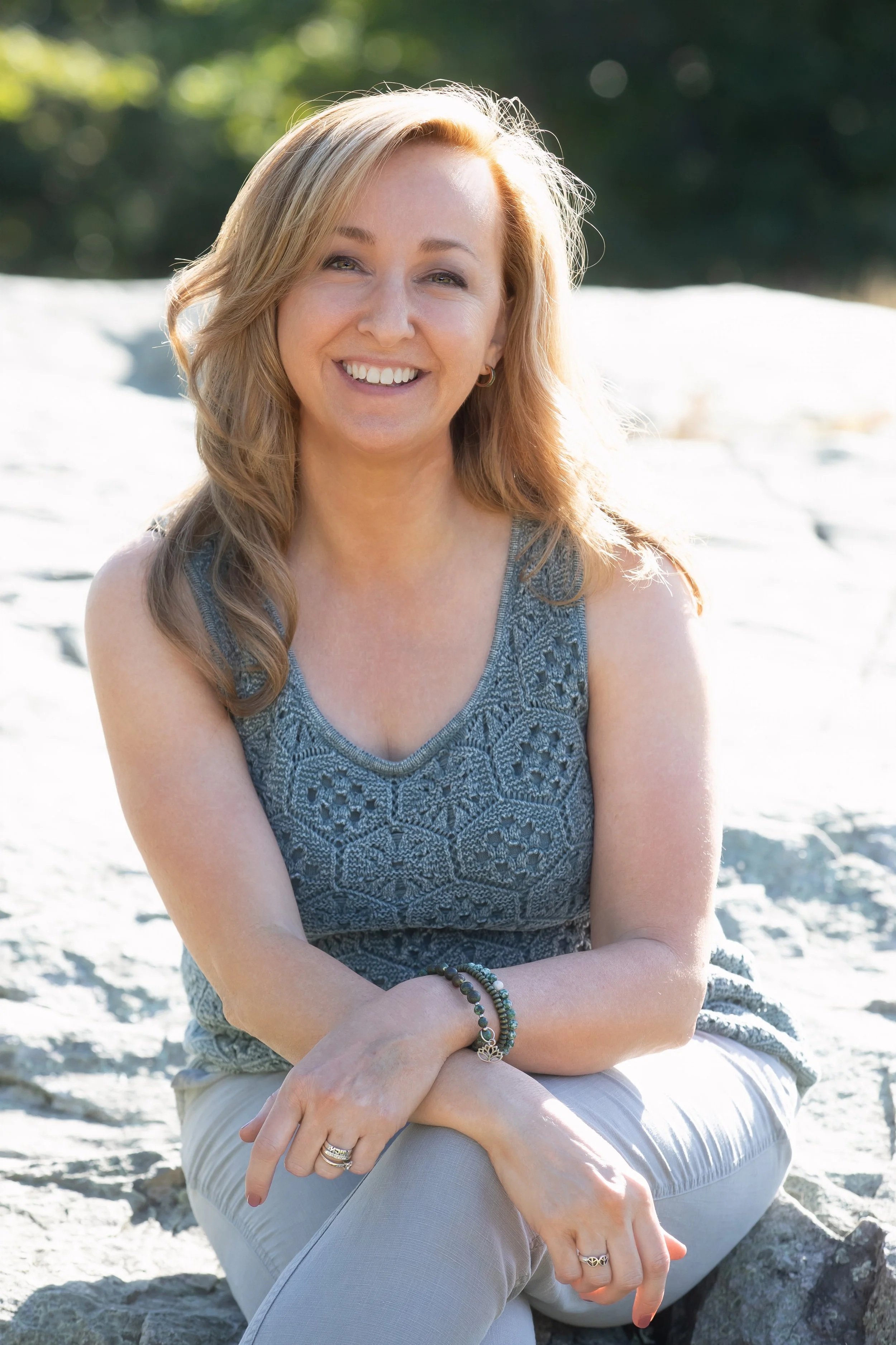 A woman with light brown, wavy hair smiling, sitting outdoors on a rocky surface, wearing a sleeveless gray patterned top and light-colored pants, with jewelry on her wrists and fingers, background of green trees and bright sunlight.