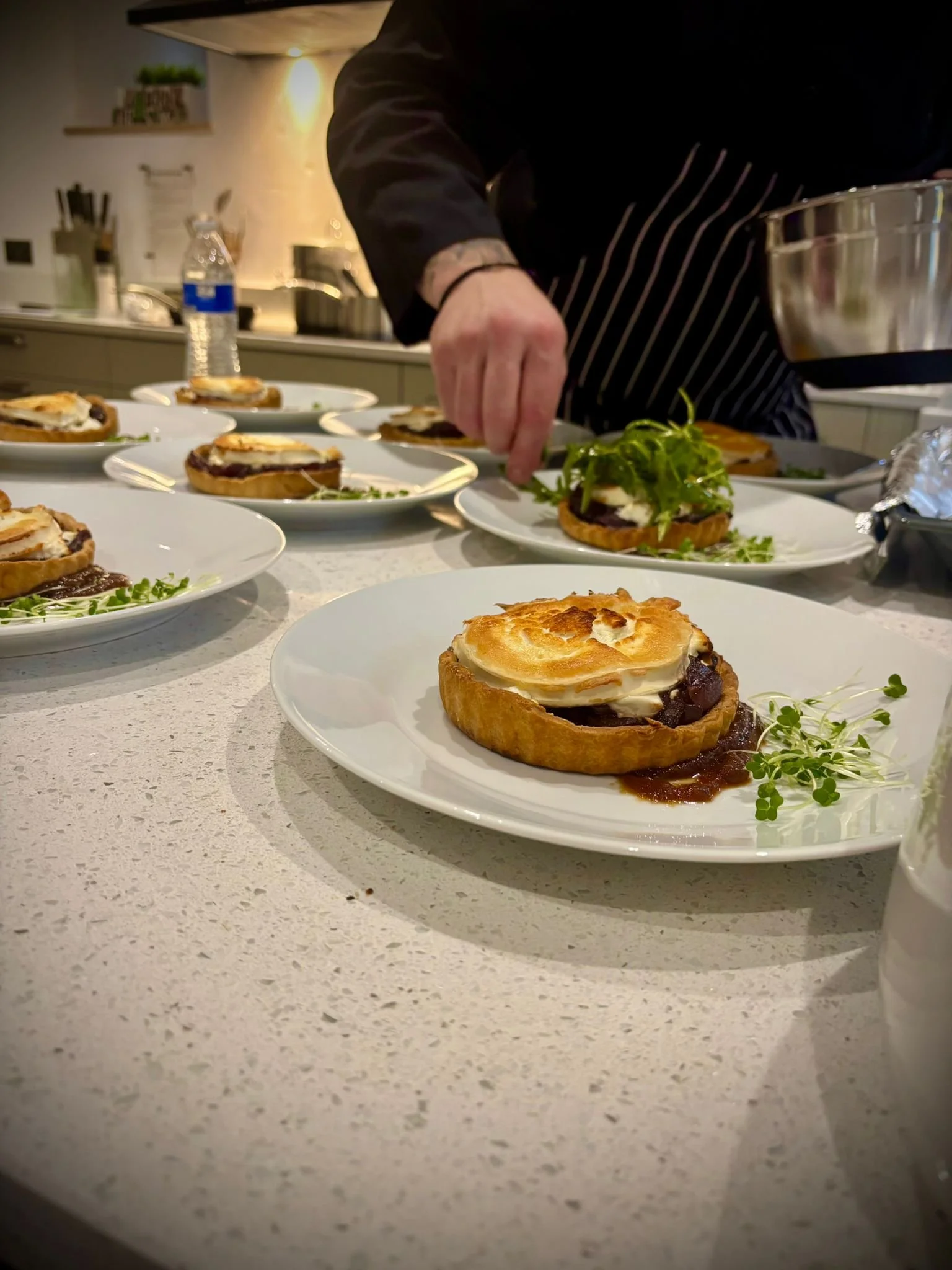 Chef arranging individual servings of savory tartlets topped with browned meringue and garnished with microgreens on a white countertop.
