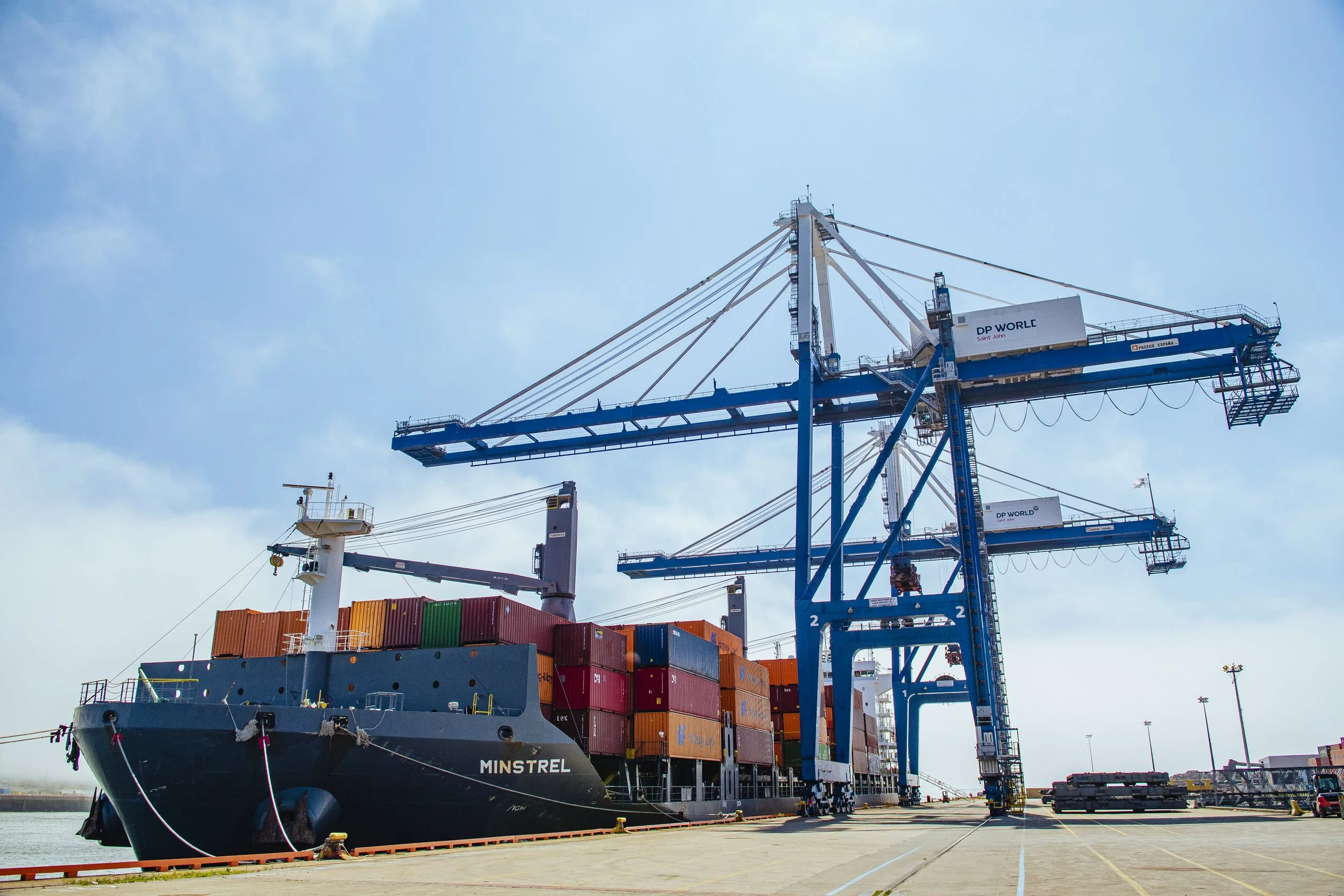 A large cargo ship named Minstrel docked at the Saint John Industrial Park port with multiple shipping containers stacked on it and three large blue cranes loading or unloading cargo.