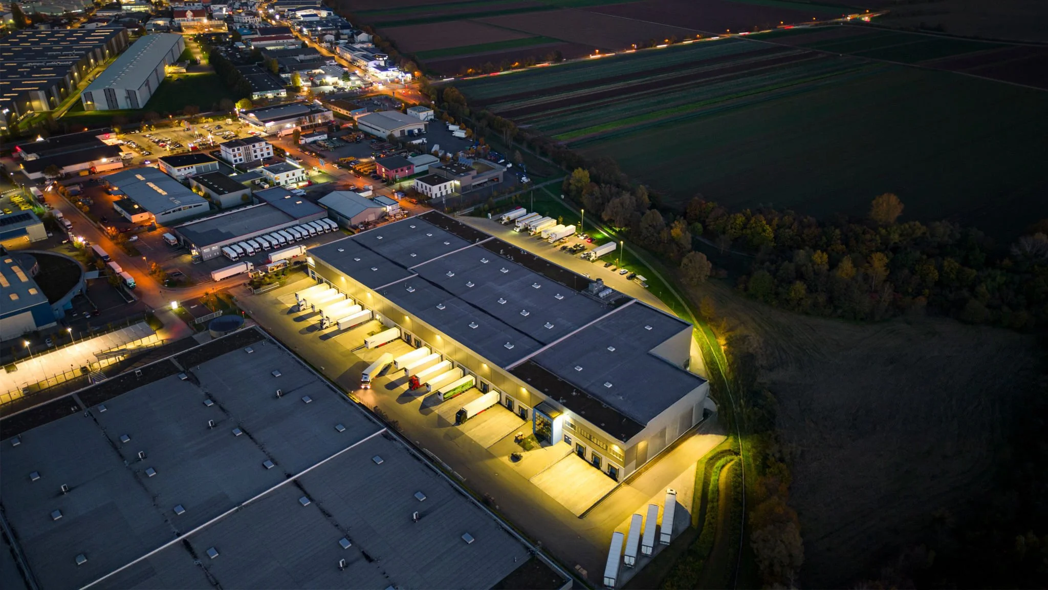 Aerial night view of an industrial complex with warehouses and loading docks, surrounded by roads and adjacent to fields, illuminated by outdoor lighting.