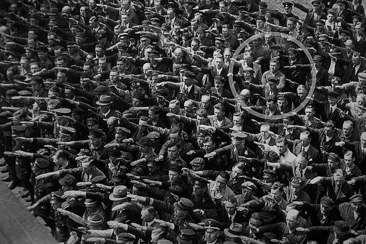Historic photograph of a crowd performing the Nazi salute, with a single man standing with arms crossed in defiance, highlighted among the group