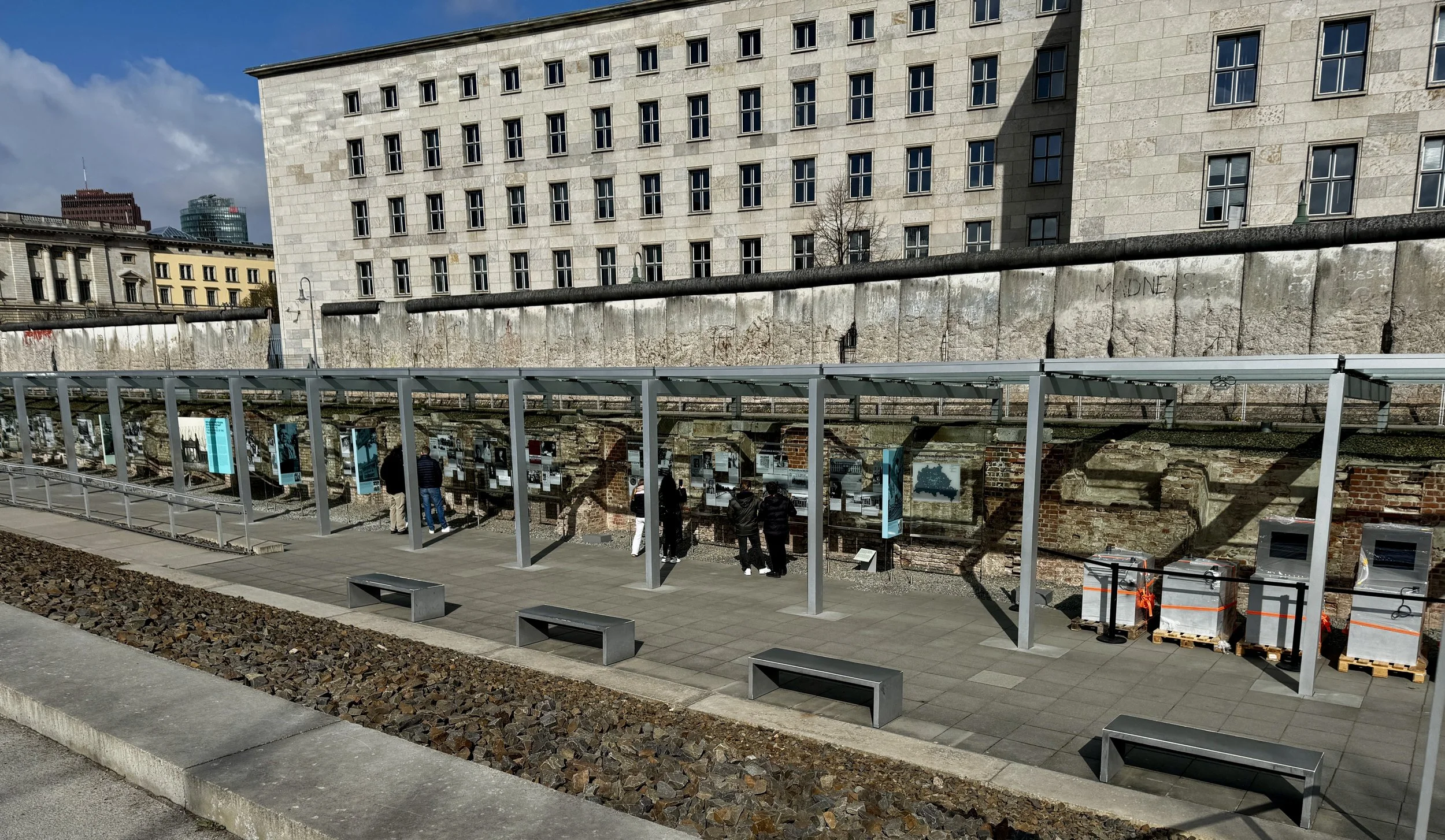 Outdoor exhibit at the Topography of Terror museum in Berlin, with preserved Berlin Wall sections and historical displays under a covered walkway.