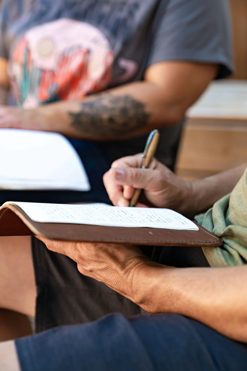 Close-up of two people taking notes; one with a pen on a white notebook, and the other holding a brown leather journal with handwritten text.